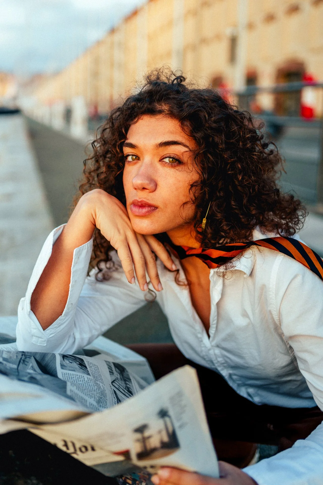 Una donna con capelli ricci scuri si appoggia al suo gomito, con uno sguardo diretto verso la fotocamera, seduta per strada con edifici sullo sfondo e un giornale aperto davanti a lei.