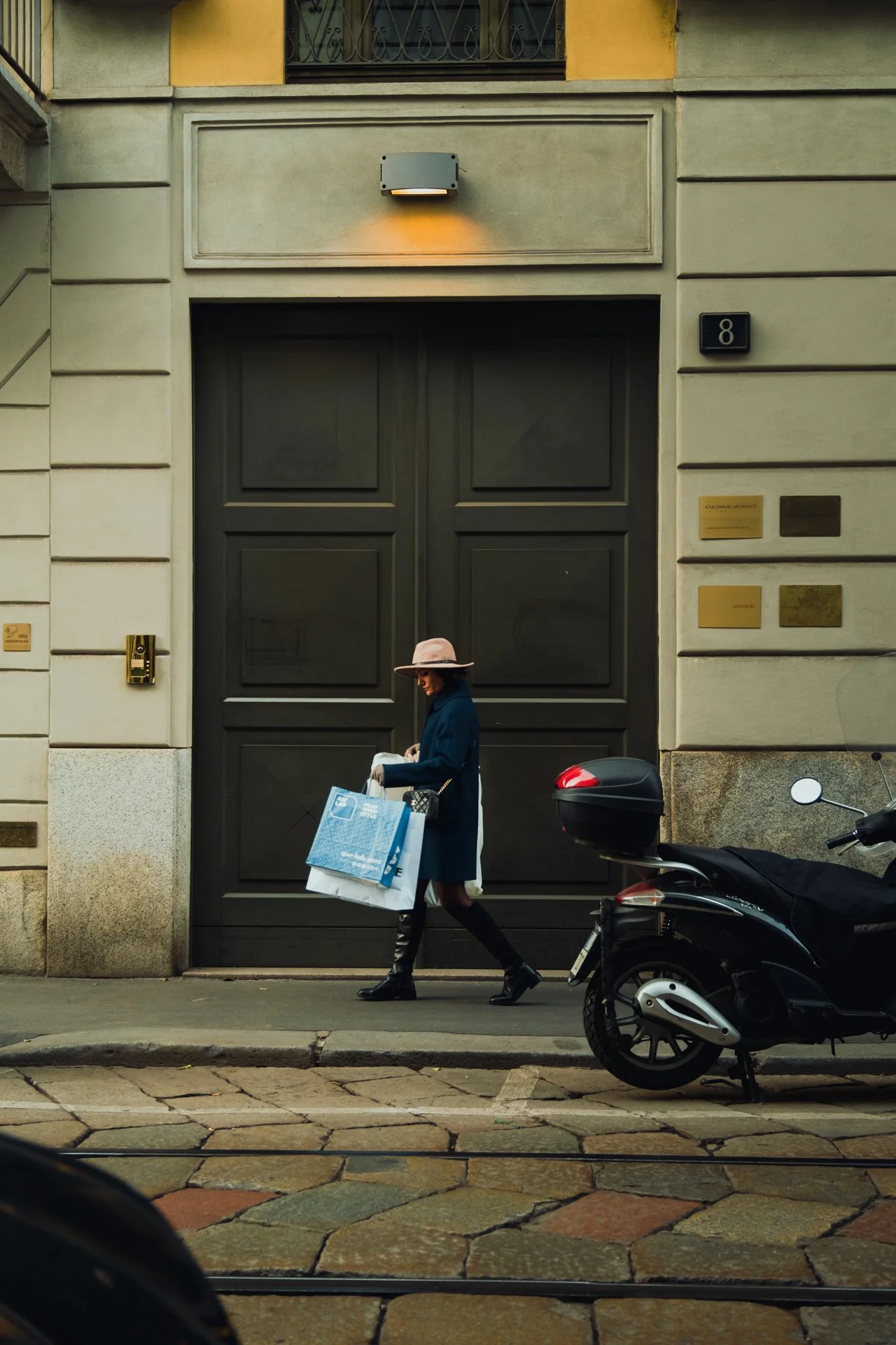 Una donna cammina lungo il marciapiede con borse, di fronte a un grande portone nero e un motorino parcheggiato.