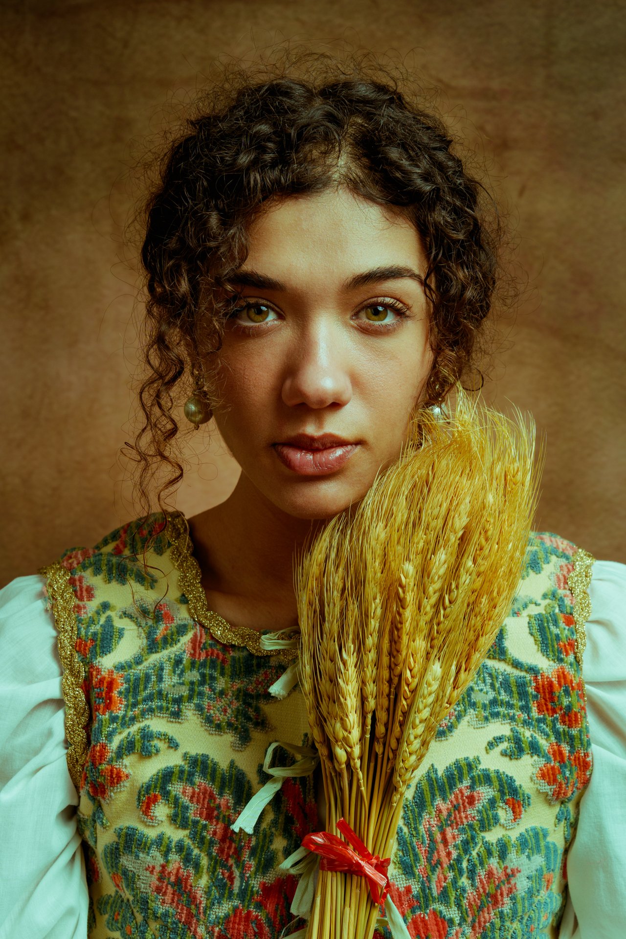 Ragazza con capelli ricci, abito tradizionale ricamato, che tiene un mazzo di spighe di grano, sfondo marrone