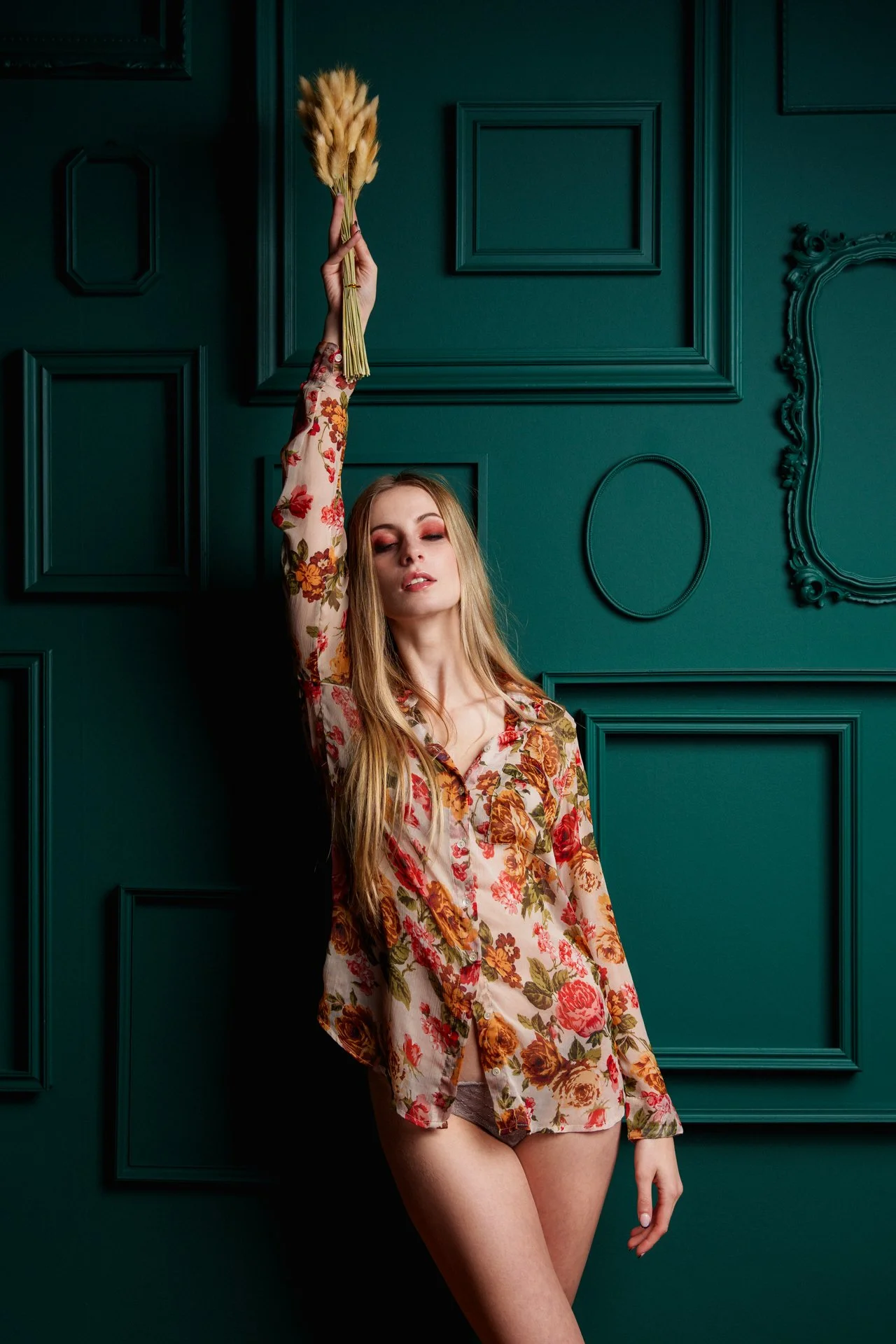 Una donna con i capelli lunghi e abbigliamento floreale sta in piedi contro una parete verde con pannelli decorativi, sollevando una mano con un mazzo di fiori secchi