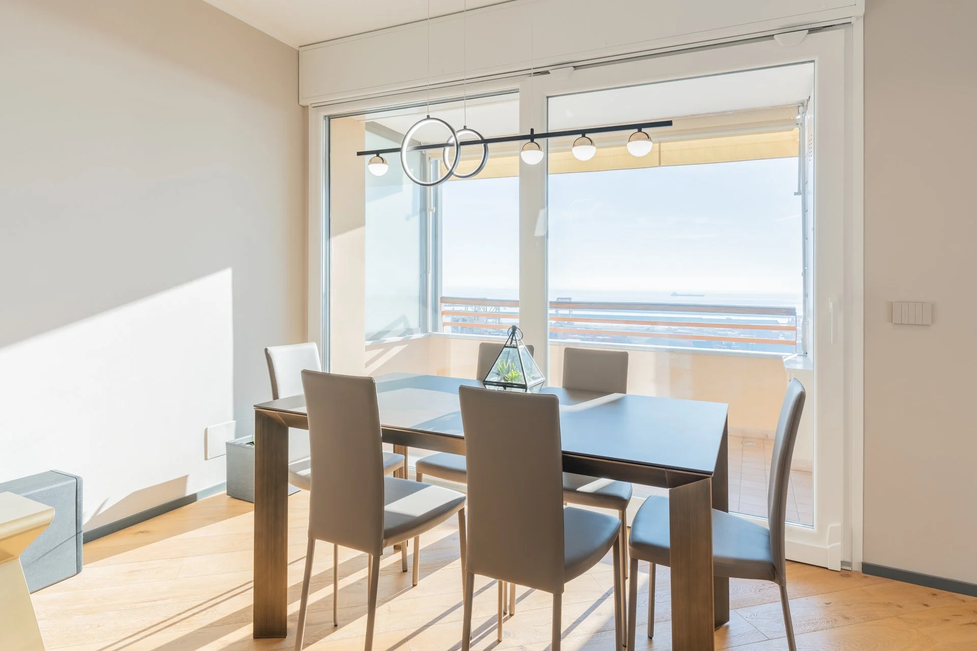 Sala da pranzo con tavolo, sei sedie e grandi vetri che danno su un balcone con vista mare.