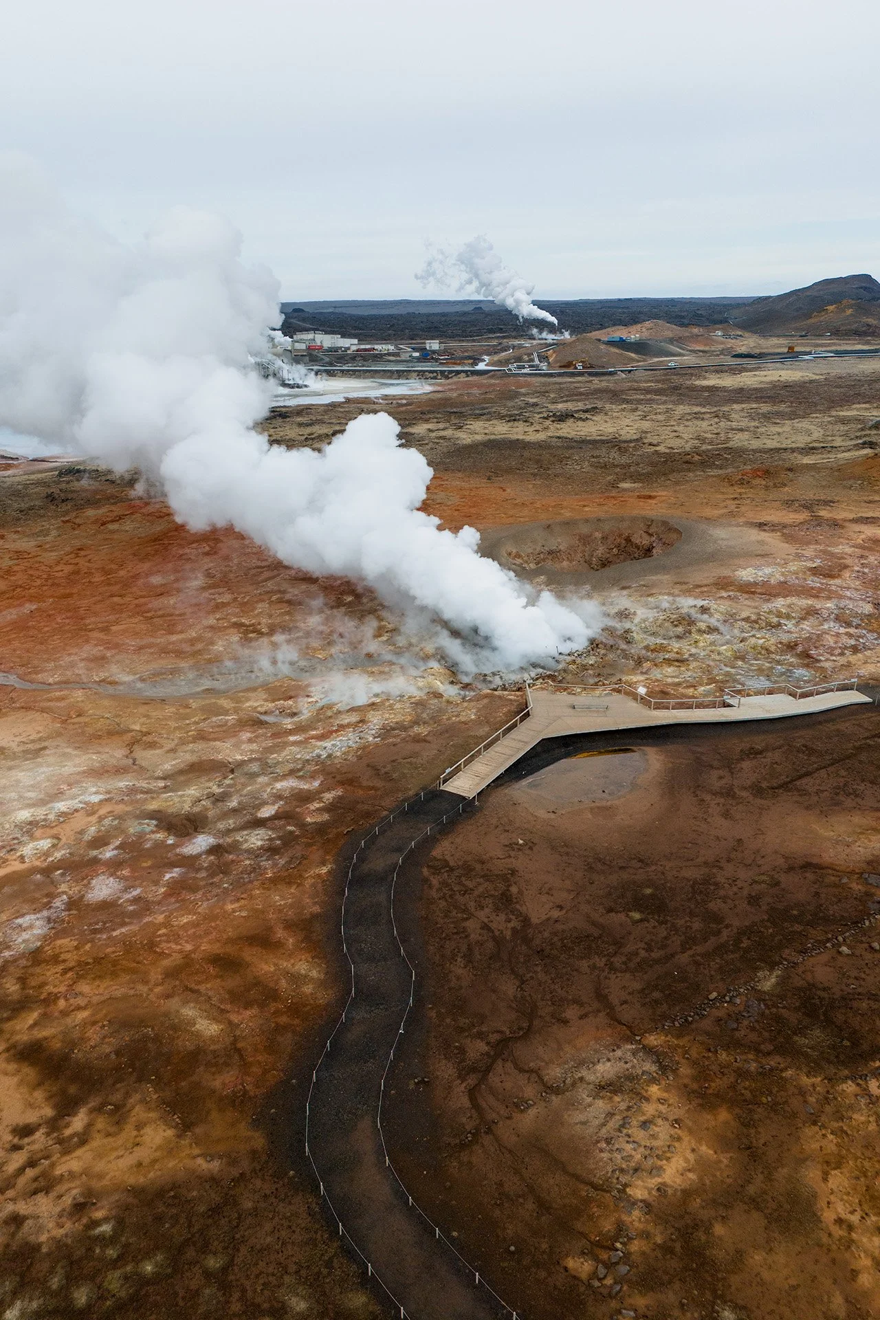 Paesaggio vulcanico con fumarole che emettono vapore bianco, sentiero di passeggiata in primo piano e terreno roccioso con tonalità marroni e arancioni. Sullo sfondo, altre fumarole e una linea di piattaforme industriali.