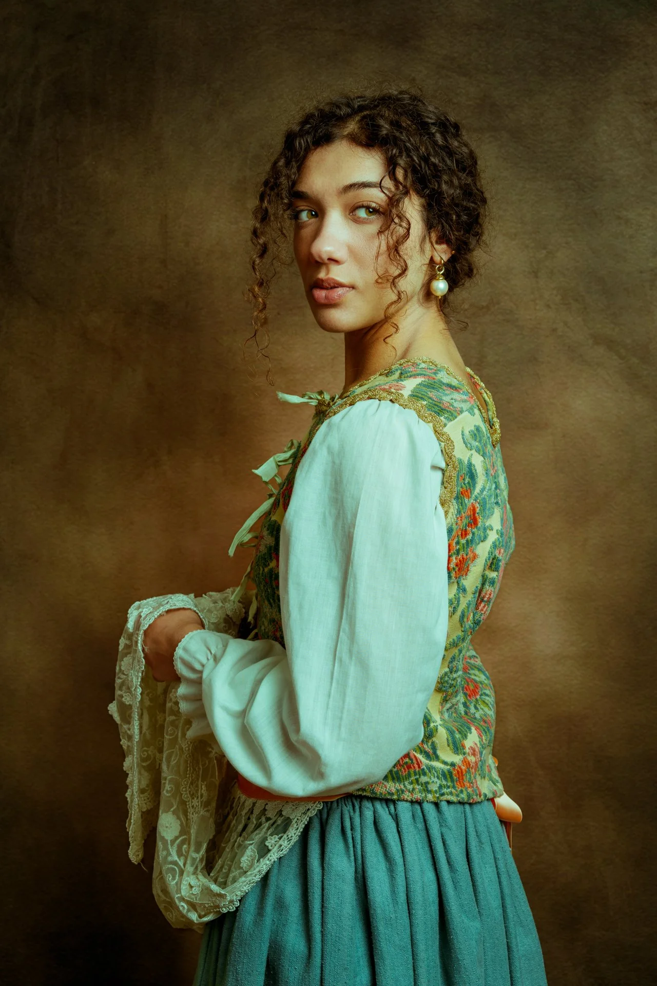 Una donna con abbigliamento tradizionale, capelli ricci, orecchini di perla, in posa su uno sfondo marrone.