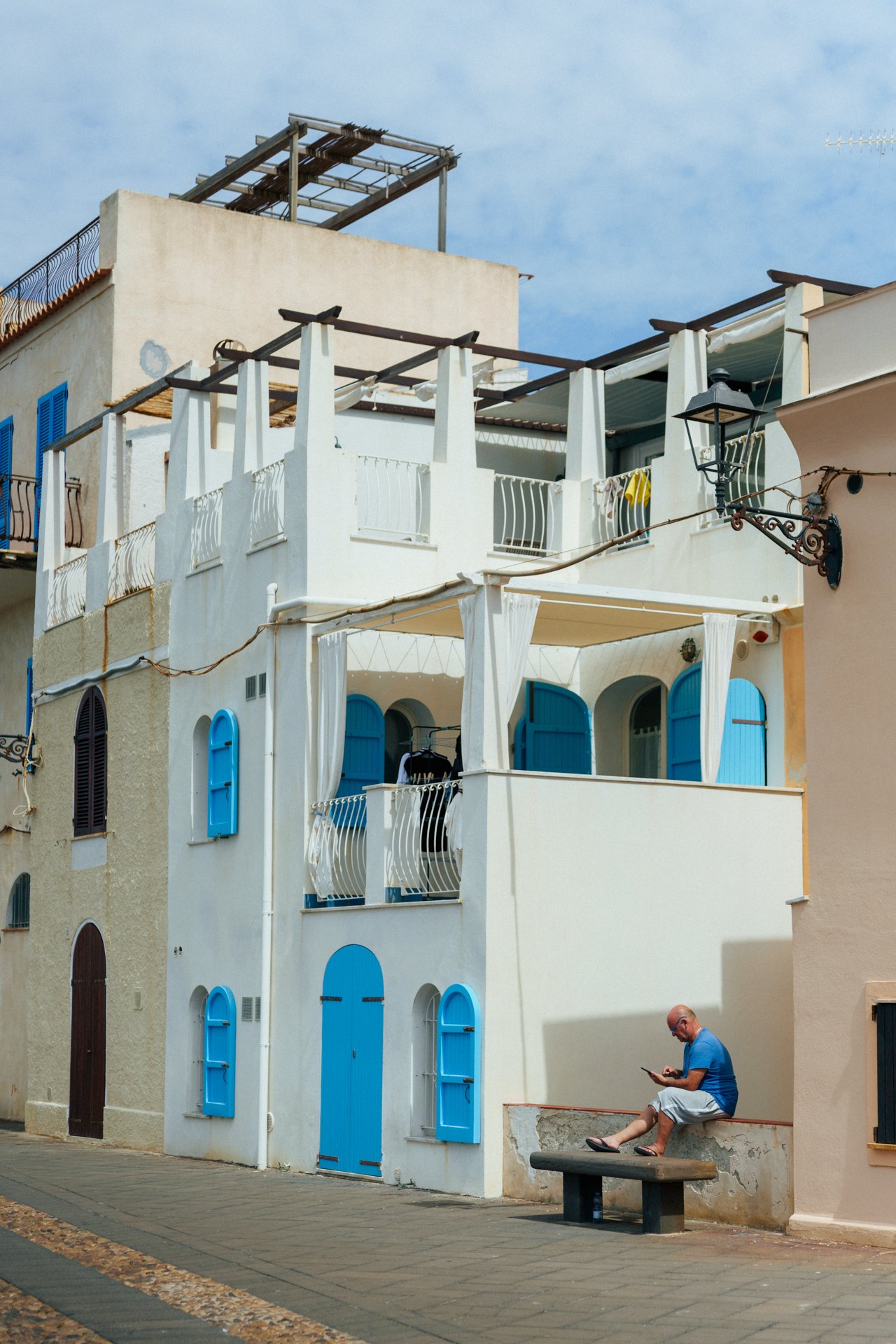 Uomo seduto su un semplice banco di pietra nella strada, intento a usare uno smartphone davanti a un edificio bianco con persiane blu, in stile mediterraneo.