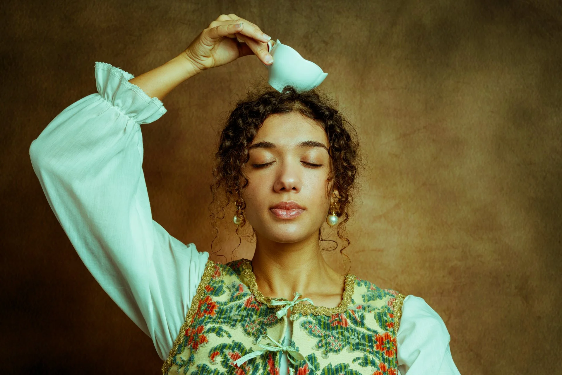 Donna con capelli riccioli e orecchini perle chiude gli occhi mentre versa acqua sulla testa.