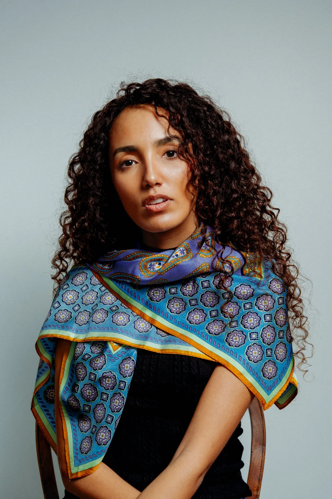Ragazza con capelli ricci, indossa una sciarpa colorata con motivi floreali, seduta su una sedia di legno, sfondo grigio.