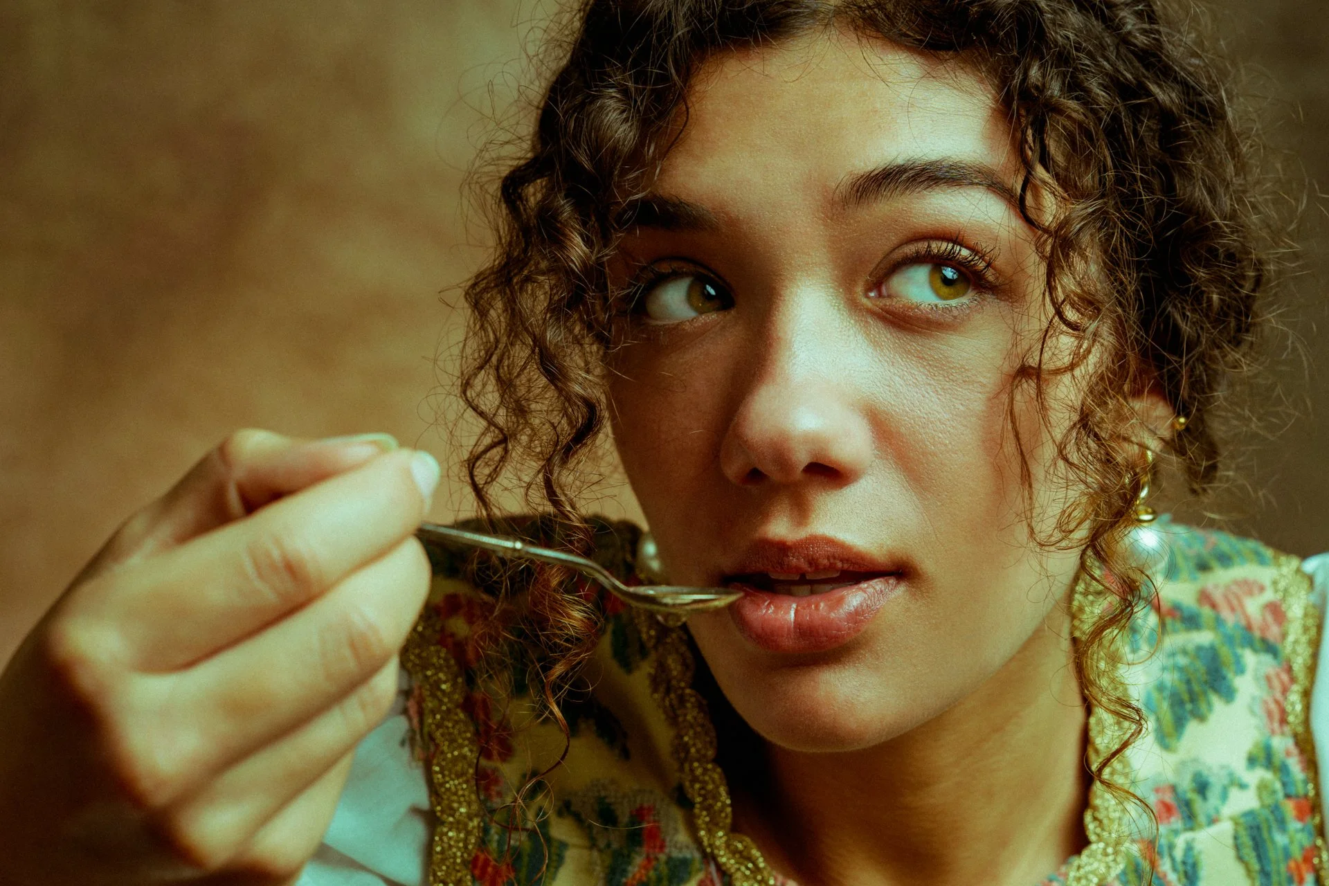 Una giovane donna con capelli riccioli e occhi verdi, che sta portando un cucchiaino vicino alle labbra, con uno sfondo sfocato e un abito colorato.