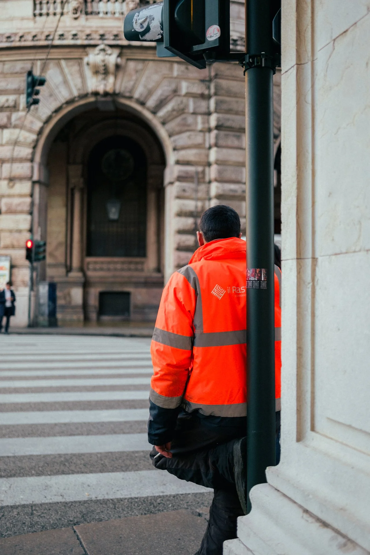 Persona in abbigliamento arancione con scritto 'Il RAS' che si appoggia a un palo in una strada di città, con un grande edificio in pietra sullo sfondo.