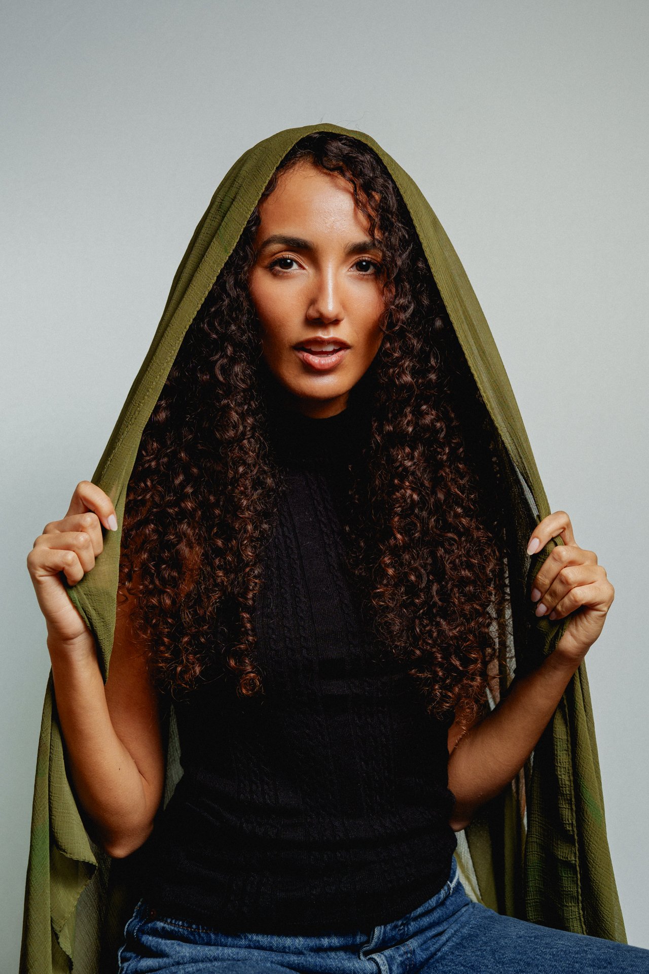 Una donna giovane con riccioli castani tiene una giacca verde sulla testa, indossa un maglione nero e jeans, sfondo grigio.