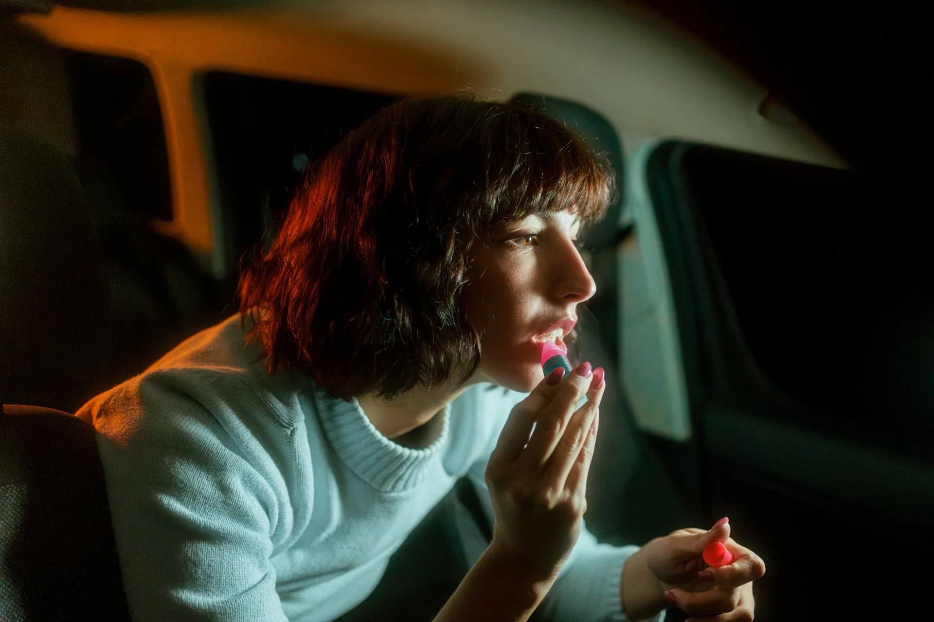 Una donna che si mette il rossetto al mattino. Sembra essere seduta sul sedile posteriore di un'auto con luci soffuse.