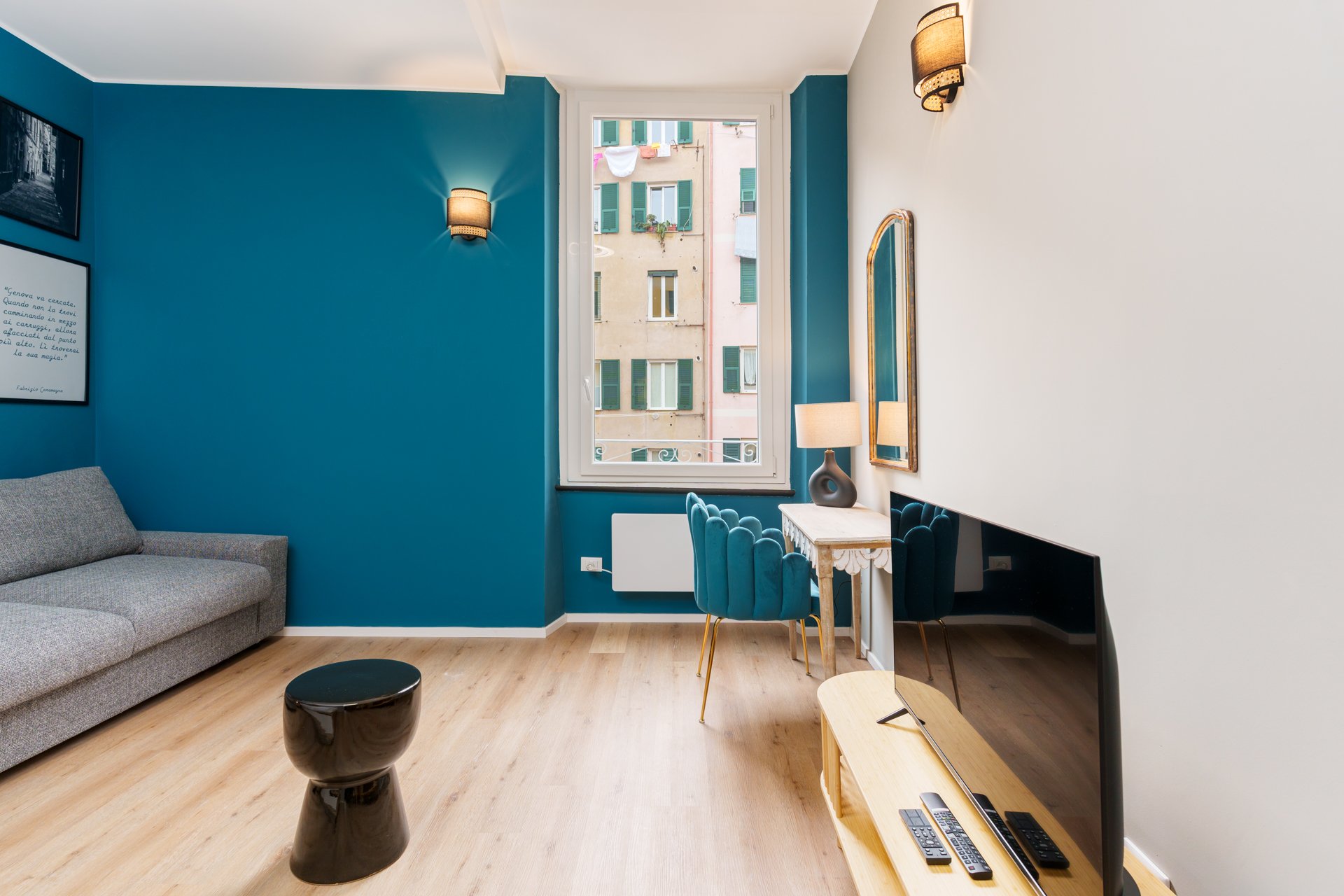 Interno di un salotto con pareti blu e bianche, un divano grigio, una televisione, una finestra con vista su un edificio, una lampada, un tavolo con sedie blu e un grande specchio, e un piccolo tavolino nero.