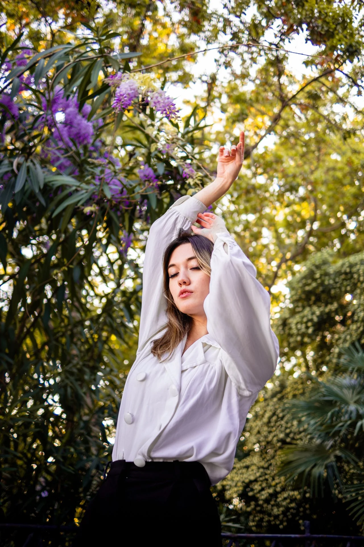 Una donna con una camicia bianca e pantaloni neri si staglia contro un background di alberi e fiori viola, con gli occhi chiusi e le braccia sollevate in una posa di serenità e stile.