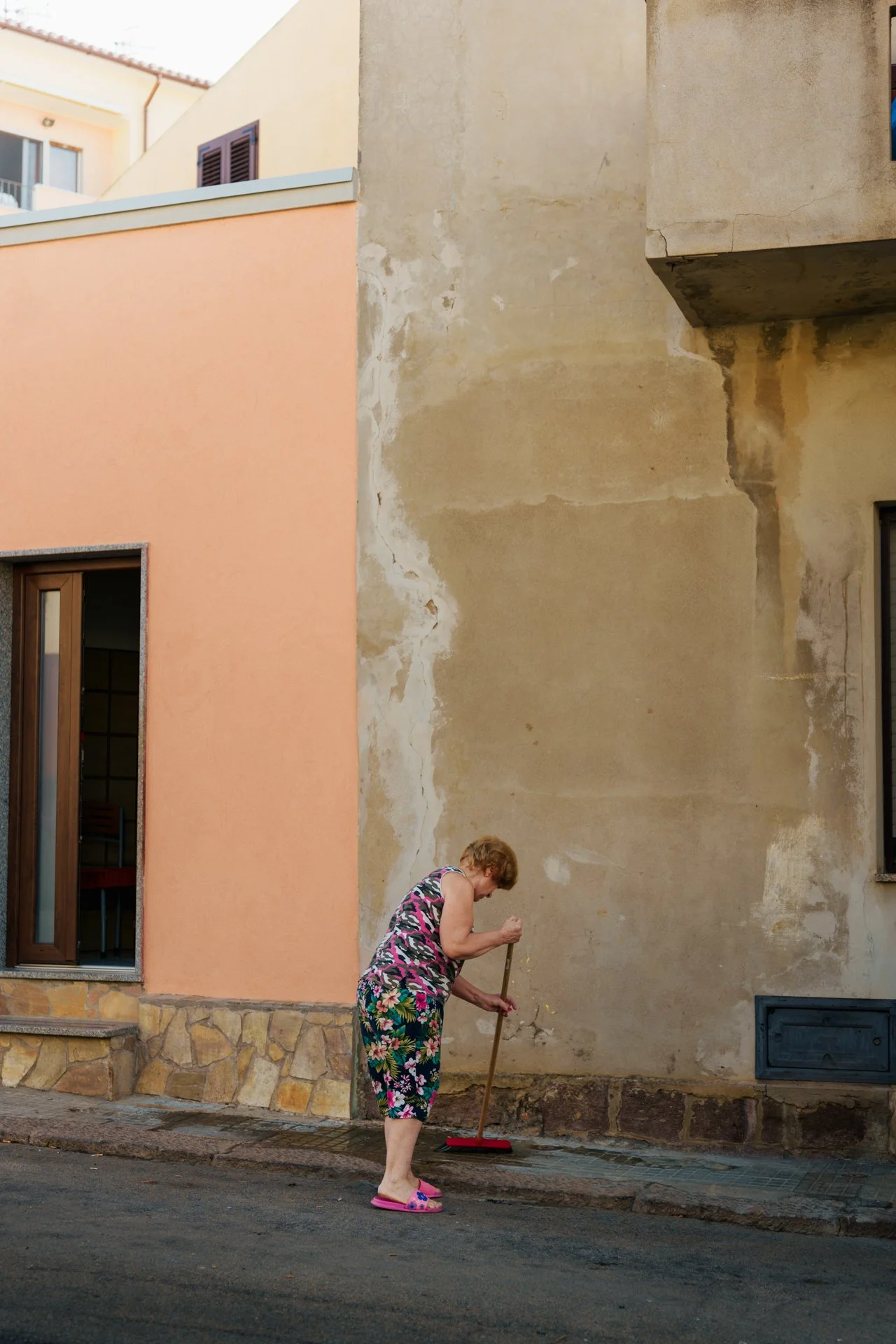 Una donna anziana che spazza il marciapiede davanti a un muro di edificio in una strada cittadina.