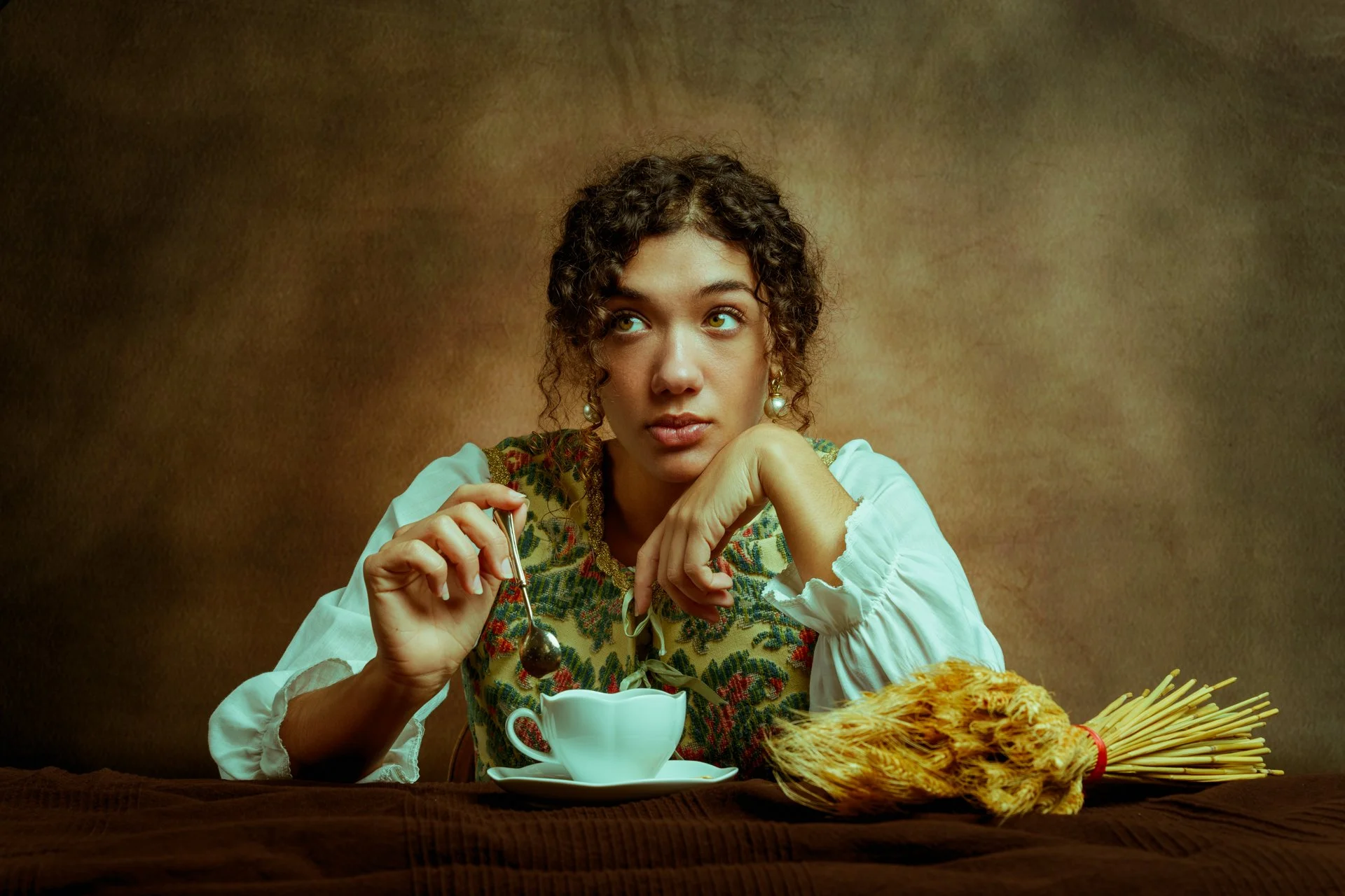 Donna con capelli riccioli corti, vestita con una camicetta bianca e un gilet decorato, seduta di fronte a una tavola con un cappuccino, un cucchiaino e un mazzo di spaghetti.