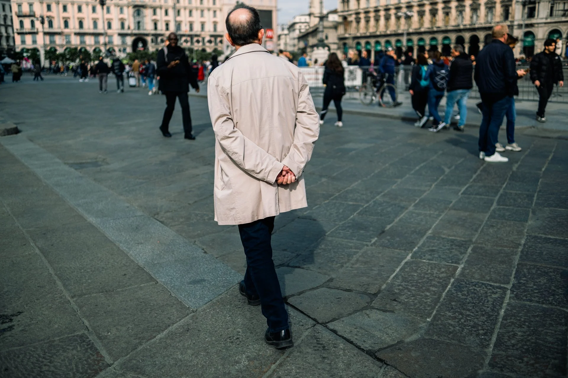 Uomo con giacca chiara che cammina in una piazza affollata con persone che camminano e parlano.