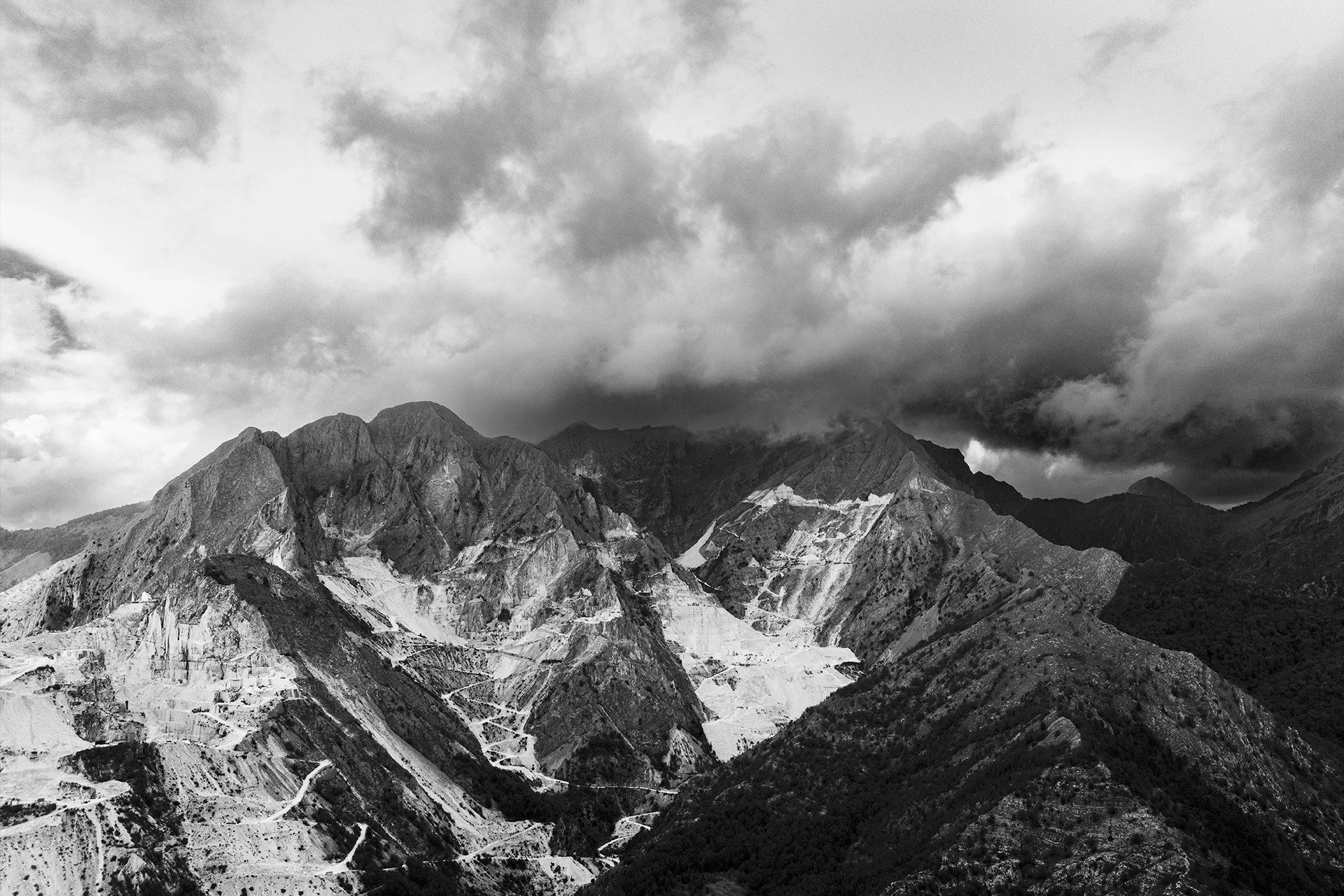 Montagne scure con nuvole dense sopra, alcune zone di scavi o terra lasciata a vista sulle pendici delle montagne.