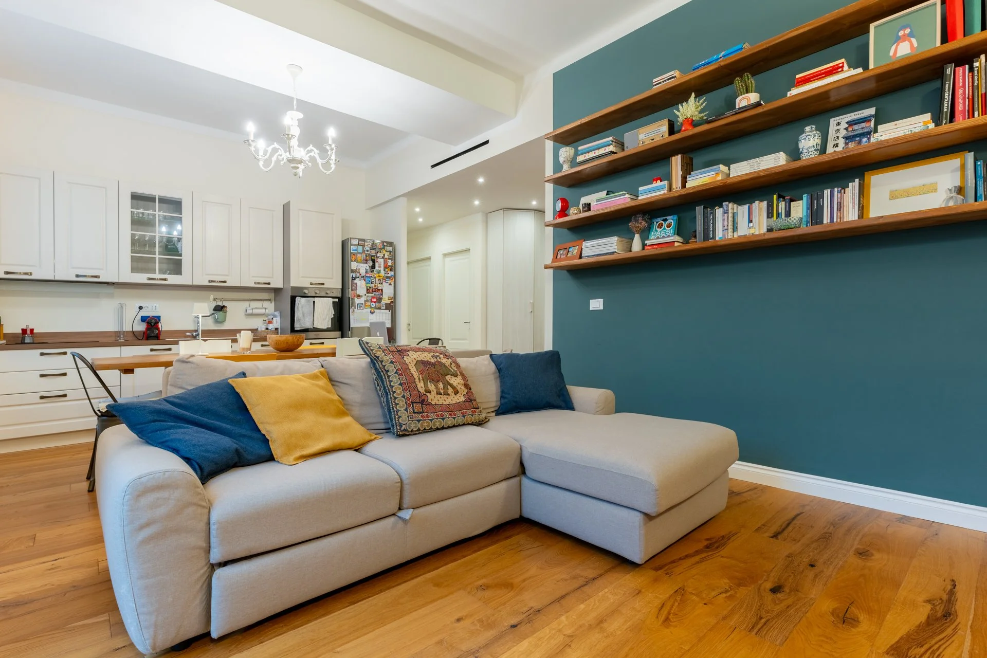 Soggiorno con divano beige, cuscini blu e giallo, libreria di legno con libri, in cucina white con elettrodomestici, pavimento di legno, lampadario bianco.