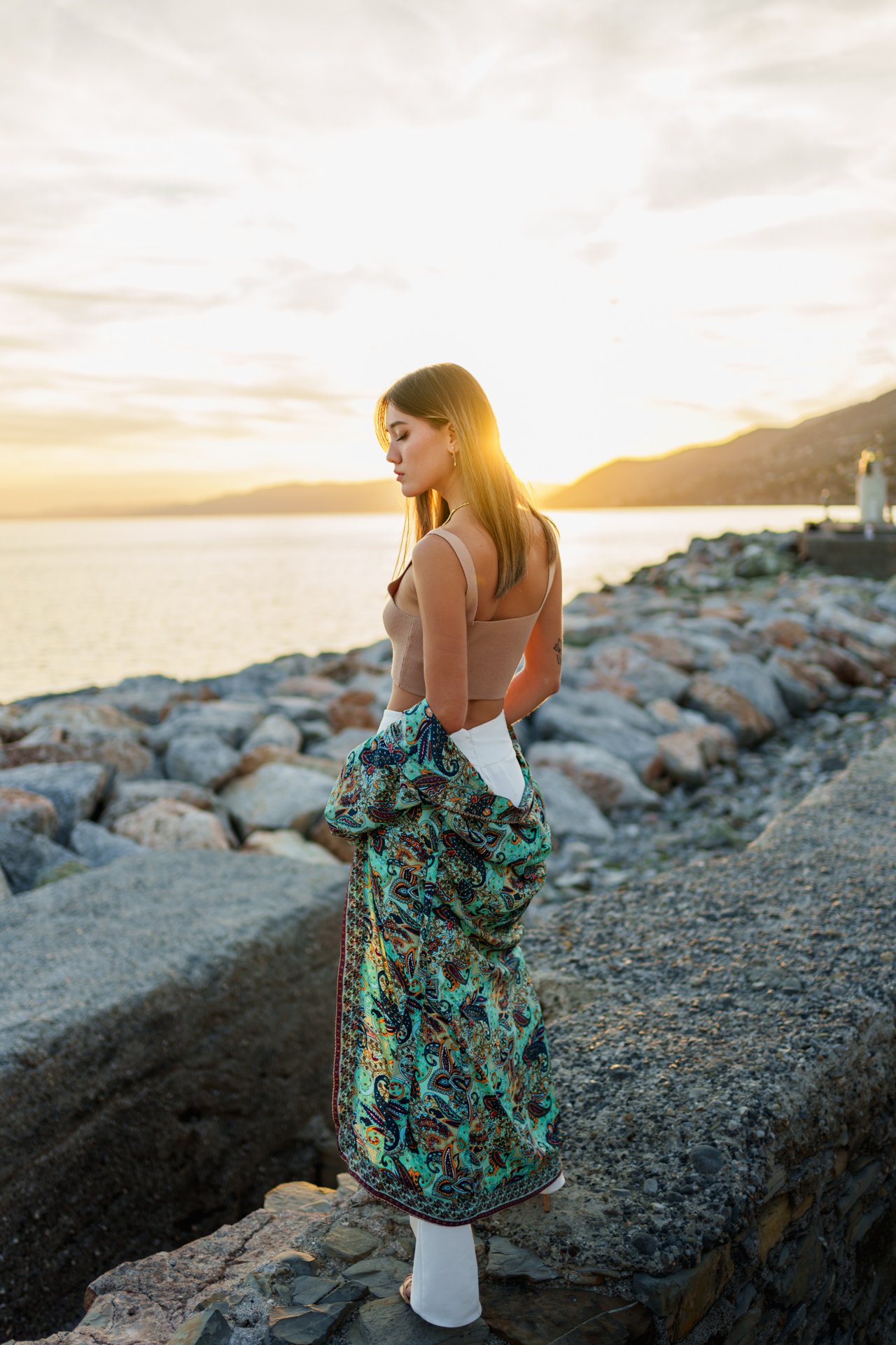 Donna che si trova sulla riva del mare durante il tramonto, indossa dei pantaloni bianchi, una gonna con un motivo colorato e una maglietta beige, con i capelli castani lisci e lunghi.