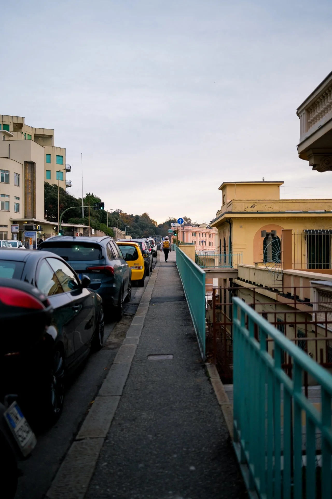 Una strada cittadina con auto parcheggiate sul lato sinistro e un marciapiede sulla destra, una persona cammina lontano con un'auto di sicurezza in fondo, edifici colorati e alberi sullo sfondo, cielo nuvoloso.