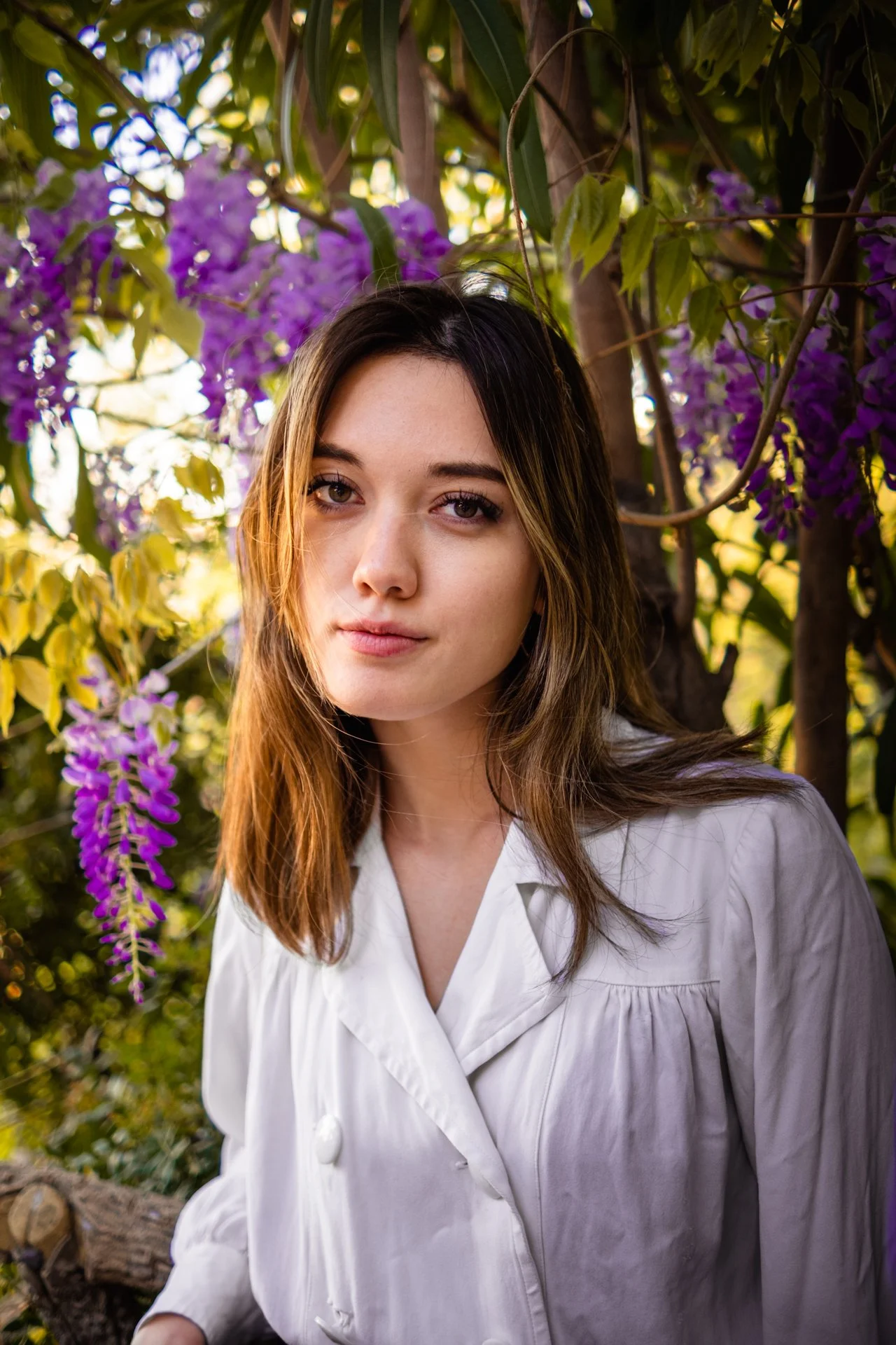 Ragazza con capelli castani chiari e vestito bianco, circondata da fiori viola e foglie verdi, in un giardino