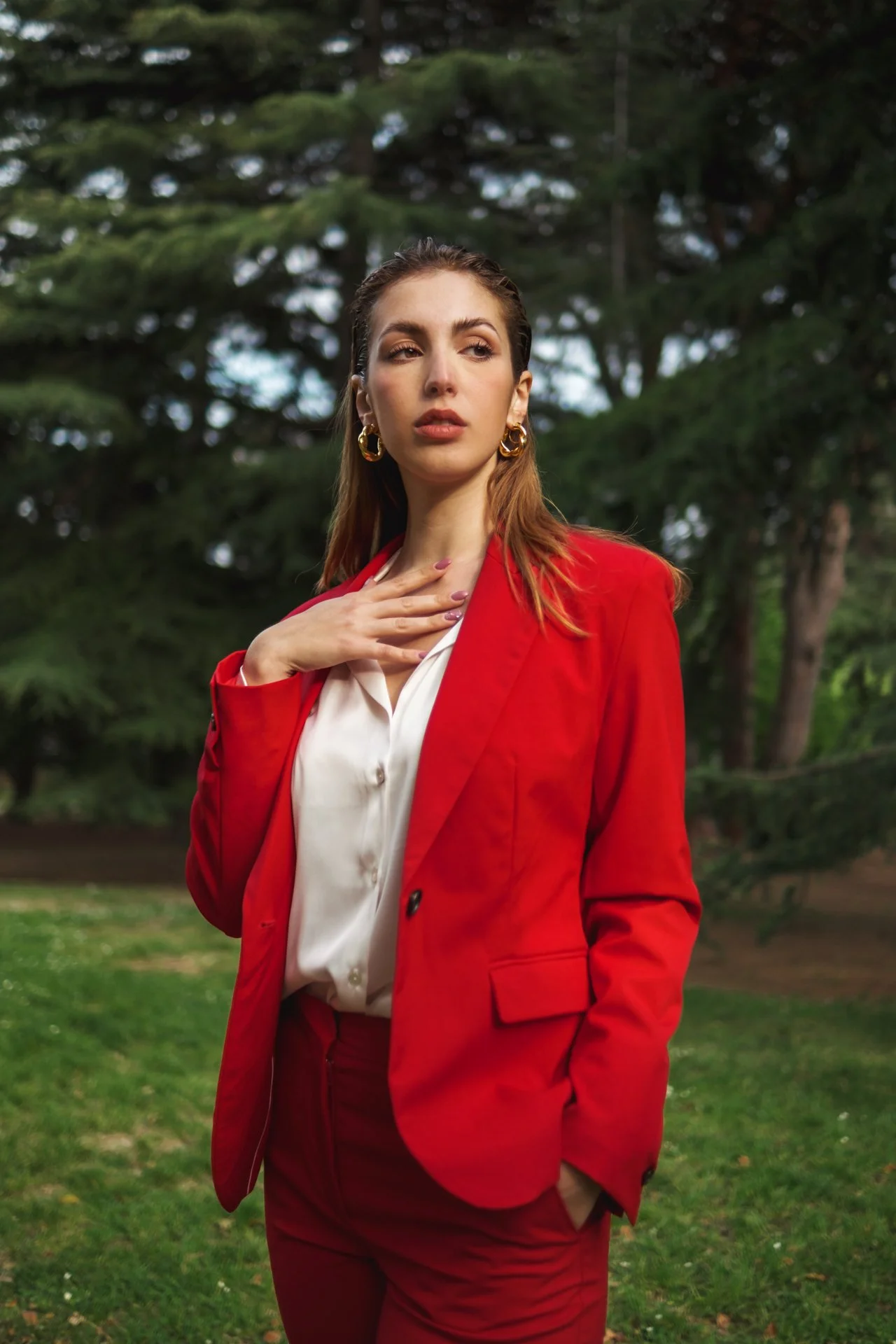 Una donna giovane con abbigliamento elegante in giacca rossa e camicia bianca si trova in un parco con alberi di sfondo.
