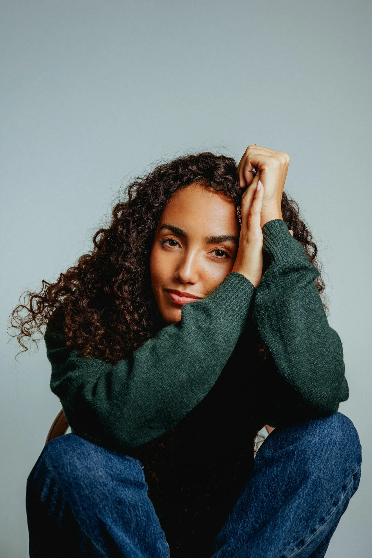 Una donna giovane con capelli ricci, intento a posare in un ritratto con sfondo neutro, indossa un maglione verde e jeans blu.
