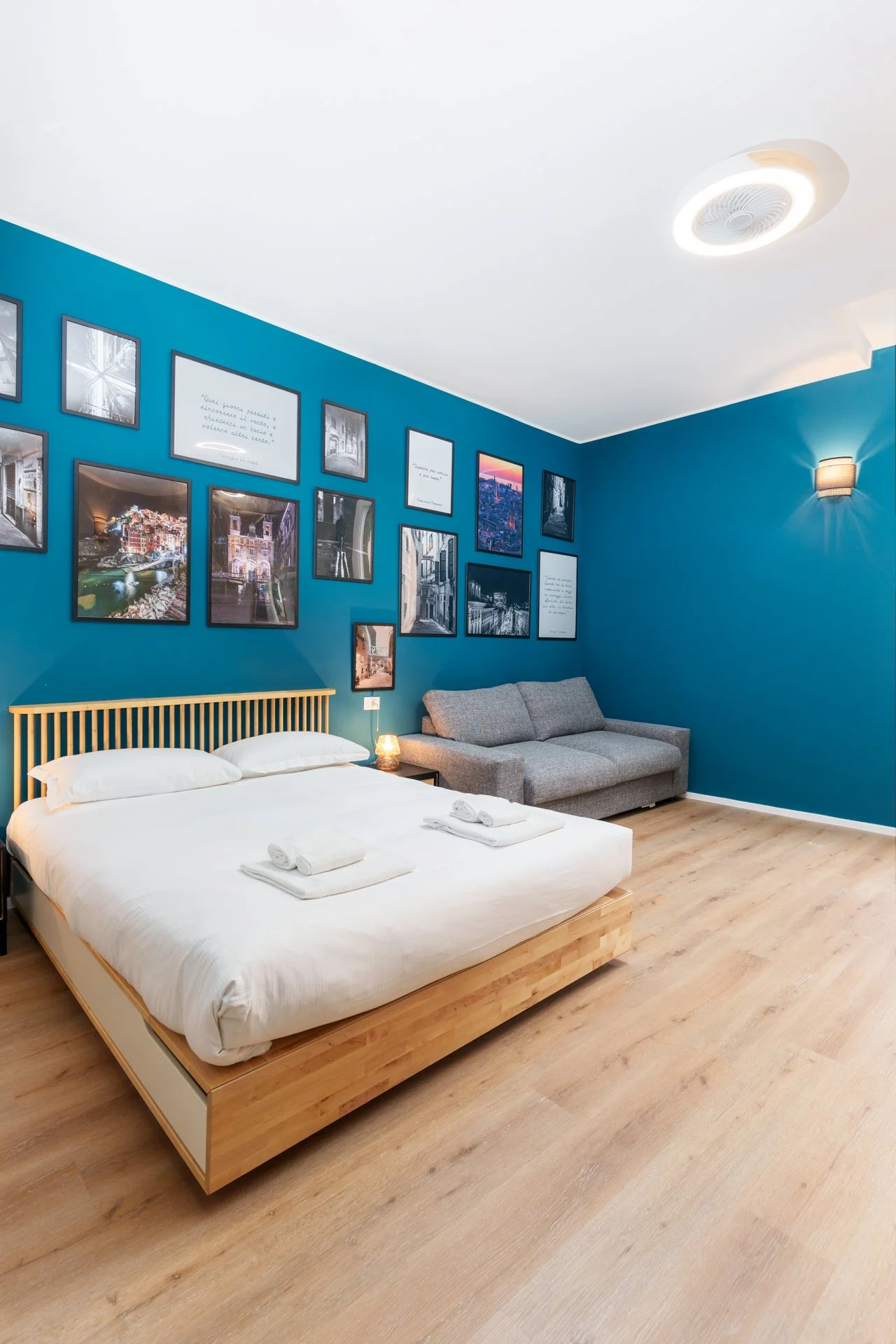 Camera da letto con parete blu, letto matrimoniale con lenzuola bianche, quadri con foto e scritte sulla parete, divano grigio, toeletta con lampada accesa e pavimento in legno chiaro.