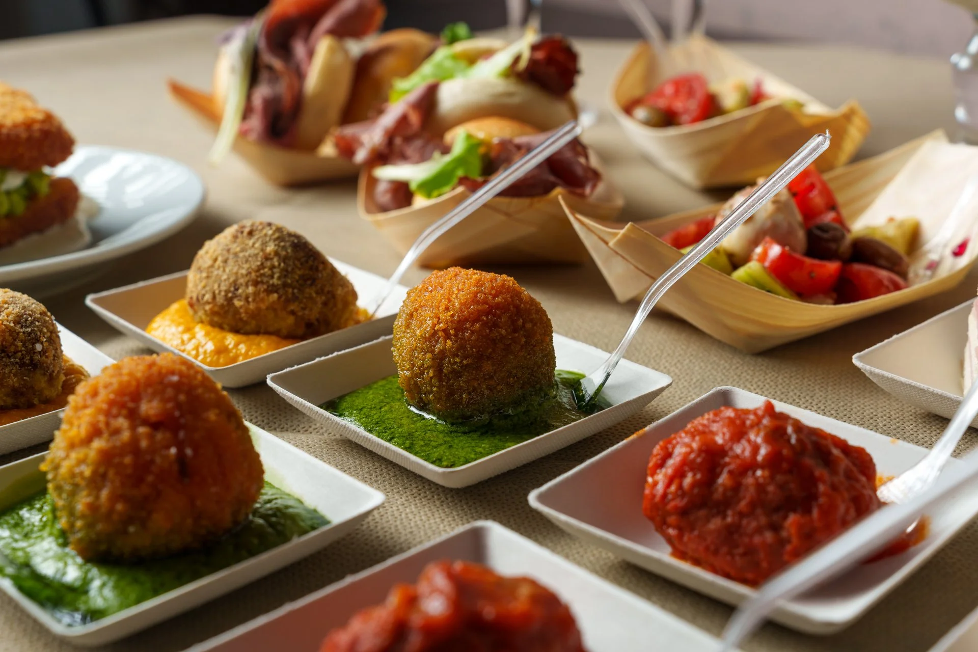 Varie polpette di diverse gusti e salse in piatti bianchi, con insalate e panini sullo sfondo