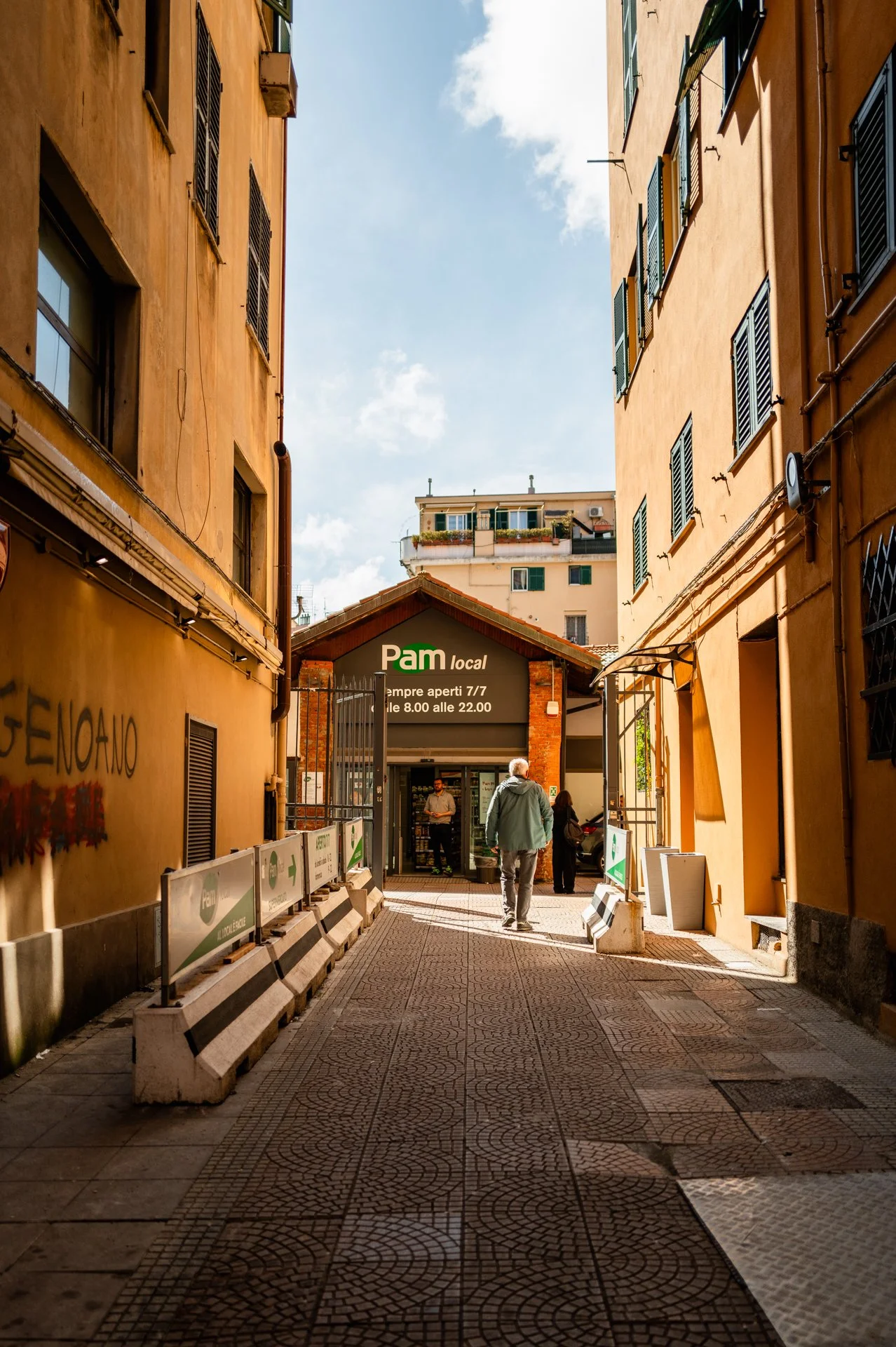 Vicolo tra due edifici gialli con negozio di alimentari chiamato Pam Local in fondo, persone che camminano verso il negozio, cielo parzialmente nuvoloso.
