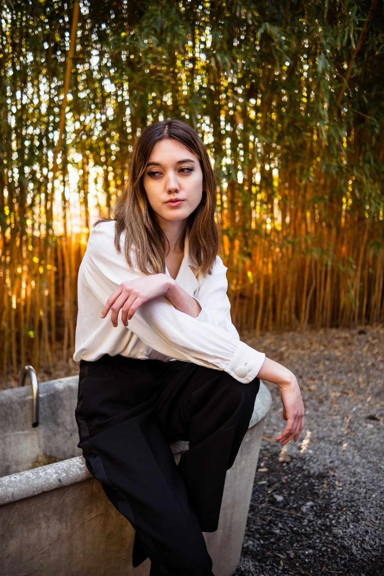 Una giovane donna con capelli marroni si siede su una fontana di pietra in un giardino con grandi cespugli di bambù, illuminata dalla luce dorata del tramonto.