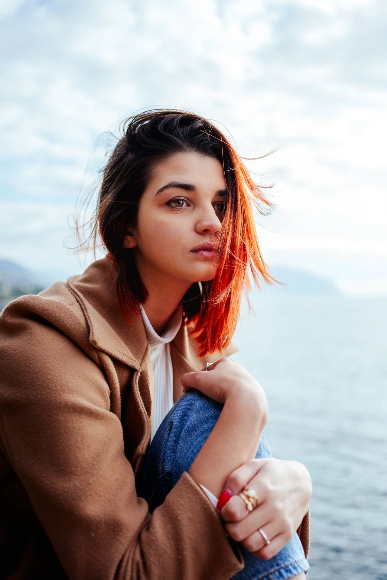 Ragazza con capelli castani e rossi seduta vicino all'acqua, guarda verso l'obiettivo, indossa una giacca marrone e ha anelli alle dita.