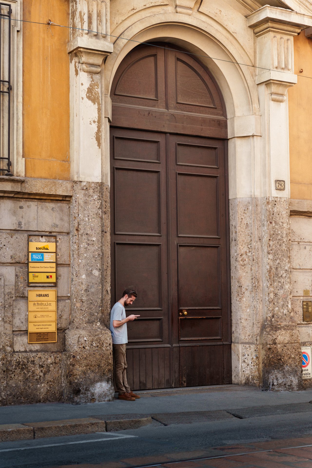 Uomo con cappotto azzurro e pantaloni marroni che usa il telefono davanti a una grande porta in legno scuro, su un edificio antico con pareti gialle e decorazioni in pietra.