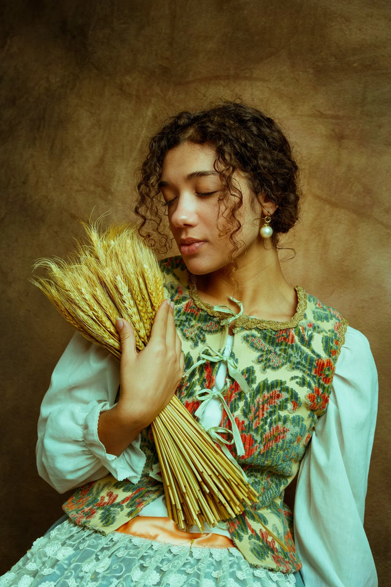Una donna con i capelli riccioli e occhi chiusi, indossa un abito tradizionale con ricami colorati e orecchini di perle, mentre tiene un mazzo di spighe di grano.