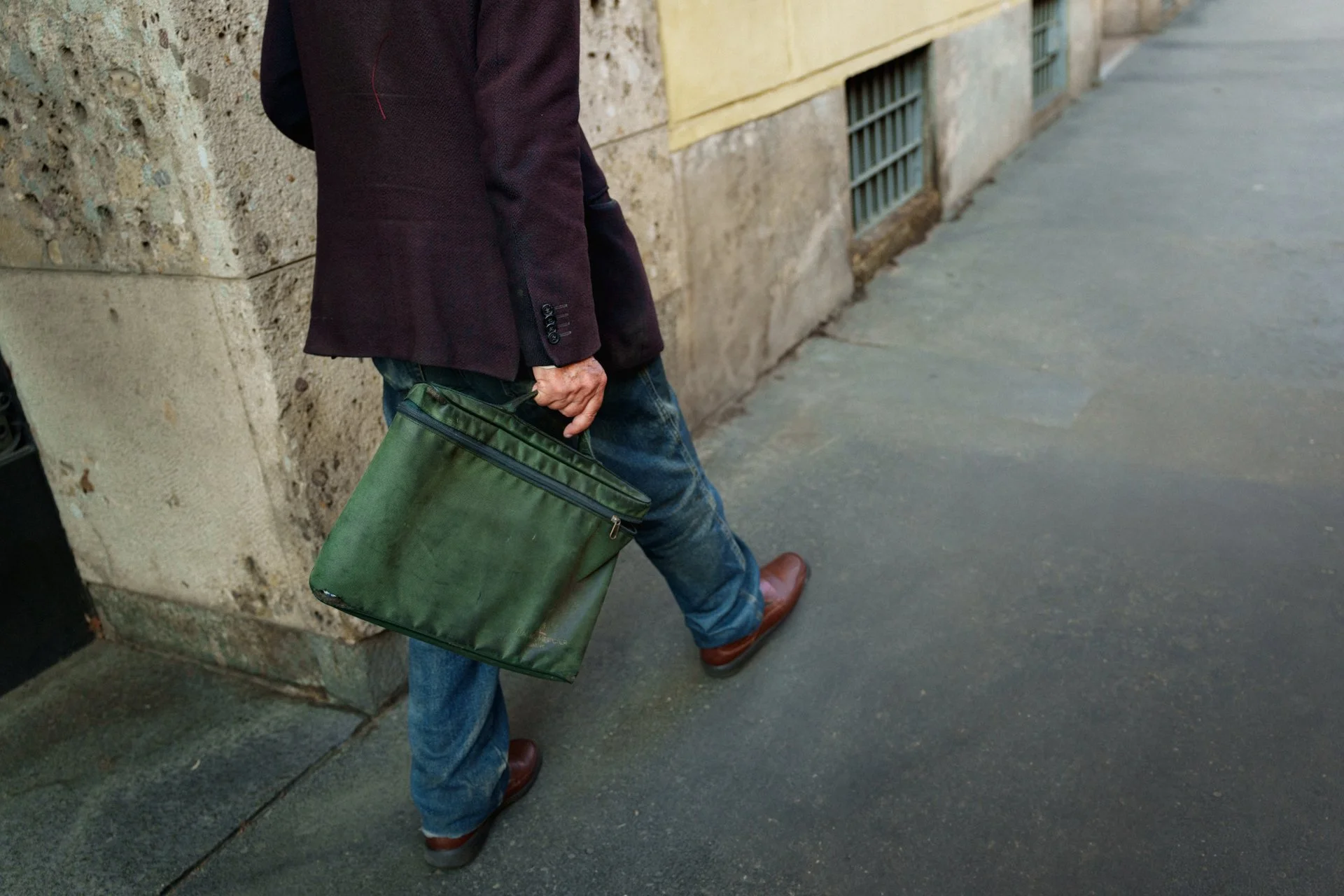 Uomo con giacca scura, jeans e scarpe di pelle, che tiene una valigetta di stoffa verde sulla mano destra, che cammina lungo un marciapiede accanto a un edificio in pietra.