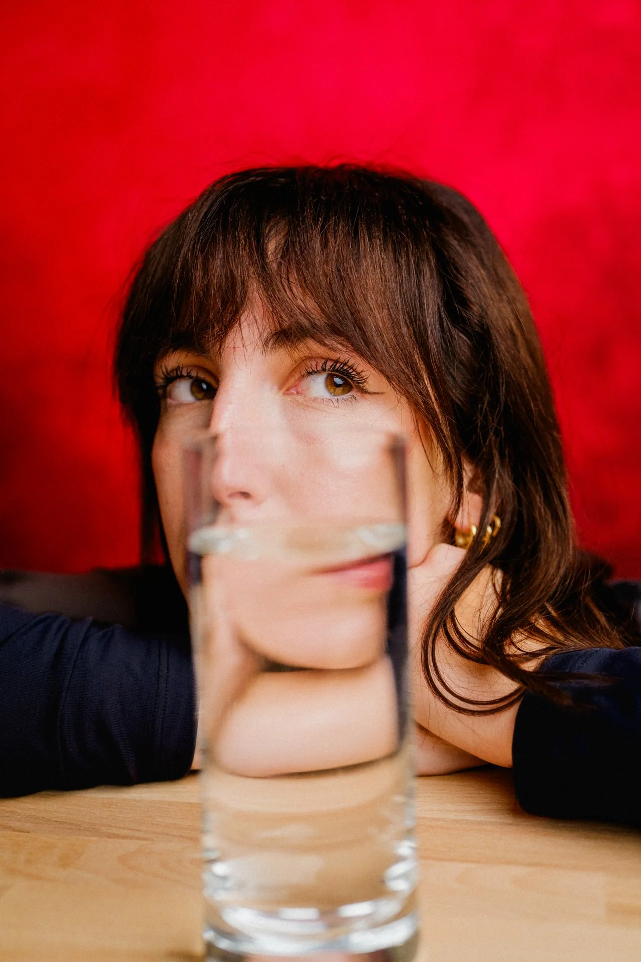 Una donna con capelli castani e occhi marroni osserva attraverso un bicchiere di acqua trasparente sulla tavola, con uno sfondo rosso.