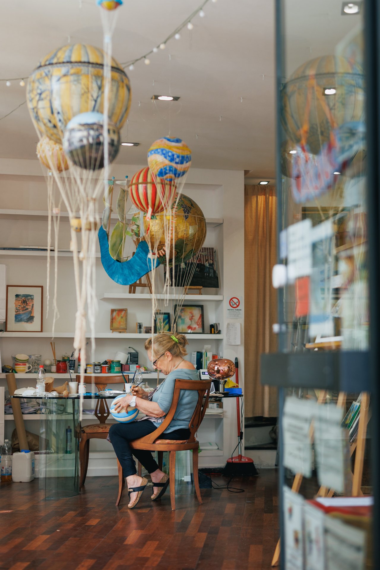 Donna anziana seduta su una sedia in un ambiente domestico o di studio, con palloncini colorati appesi al soffitto dietro di lei, che sta decorando o creando qualcosa con un pennello. L'ambiente ha scaffali con libri e oggetti decorativi, e ci sono m