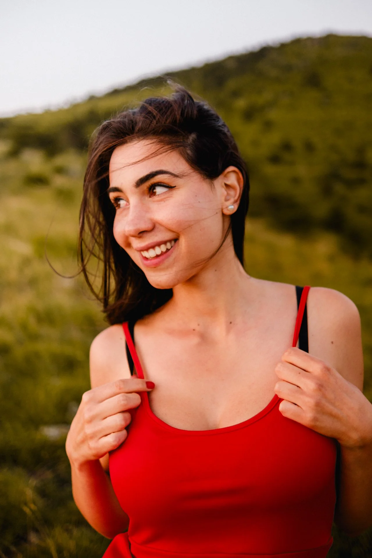 Ragazza sorridente con capelli castani, indossa un top rosso e si trova all'aperto in un paesaggio collinare verde, con un cielo chiaro di sfondo.