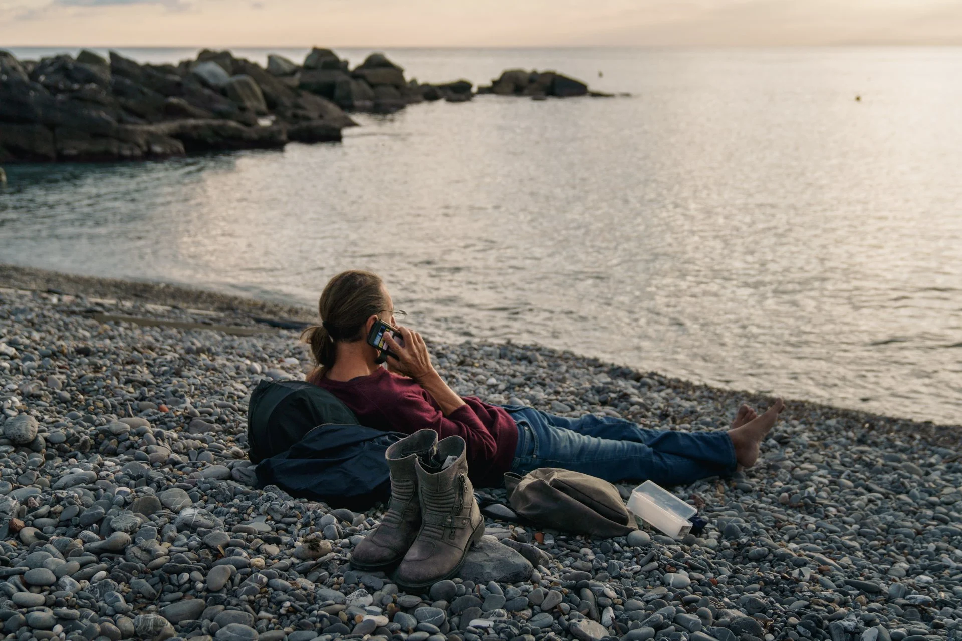 Donna che si rilassa sulla spiaggia di ciottoli durante il tramonto, mentre parla al telefono, con scarpe e oggetti personali sul terreno