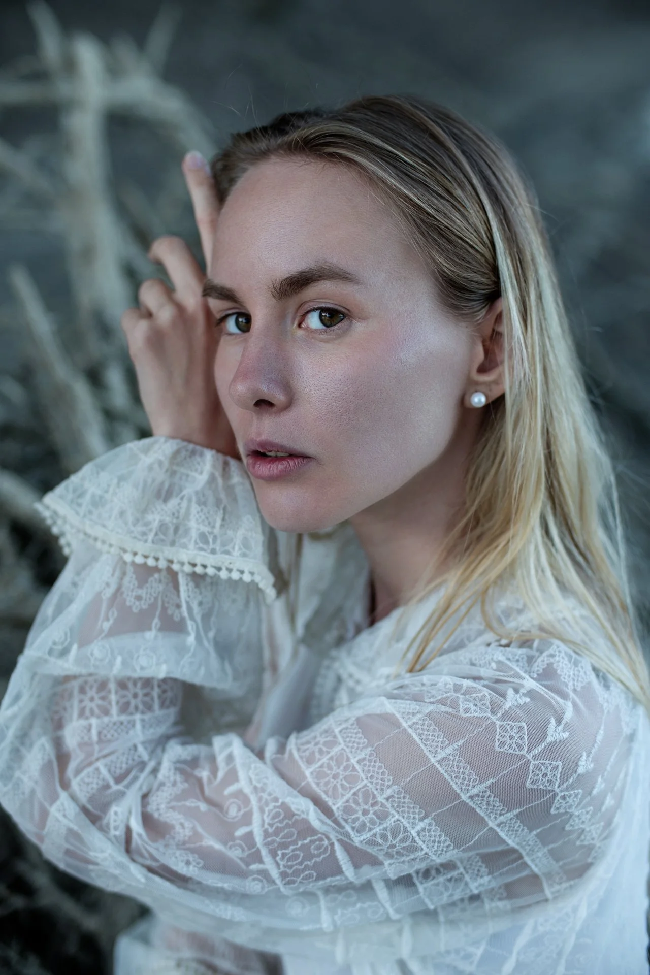 Ragazza con capelli biondi che guarda verso la fotocamera, indossa un abito di pizzo bianco con dettagli ricamati, sfondo sfocato di rami secchi.