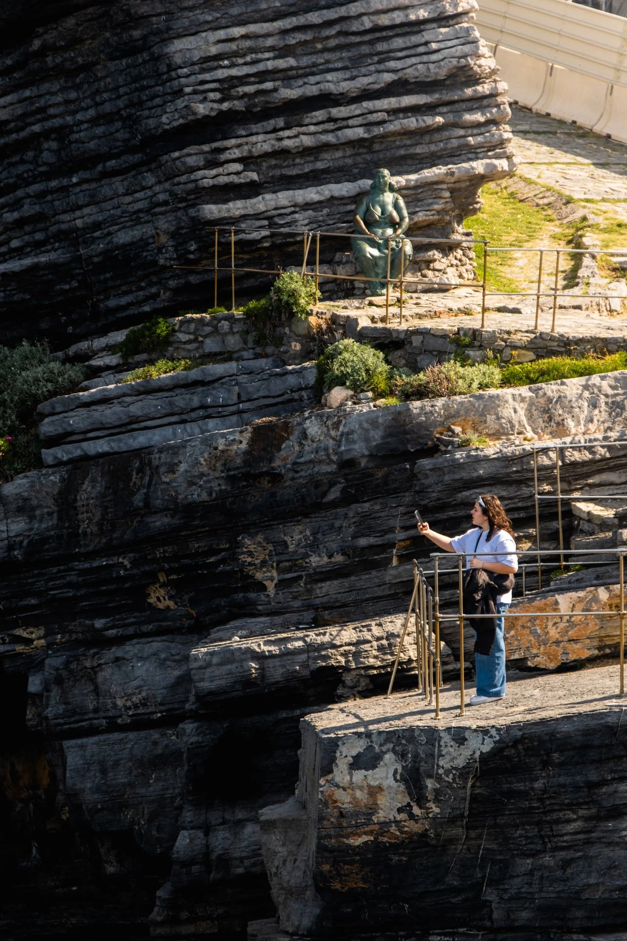 Una donna scatta una foto con il cellulare di una scultura di bronzo di un personaggio seduto, situata su una parete rocciosa di un sito storico o archeologico.