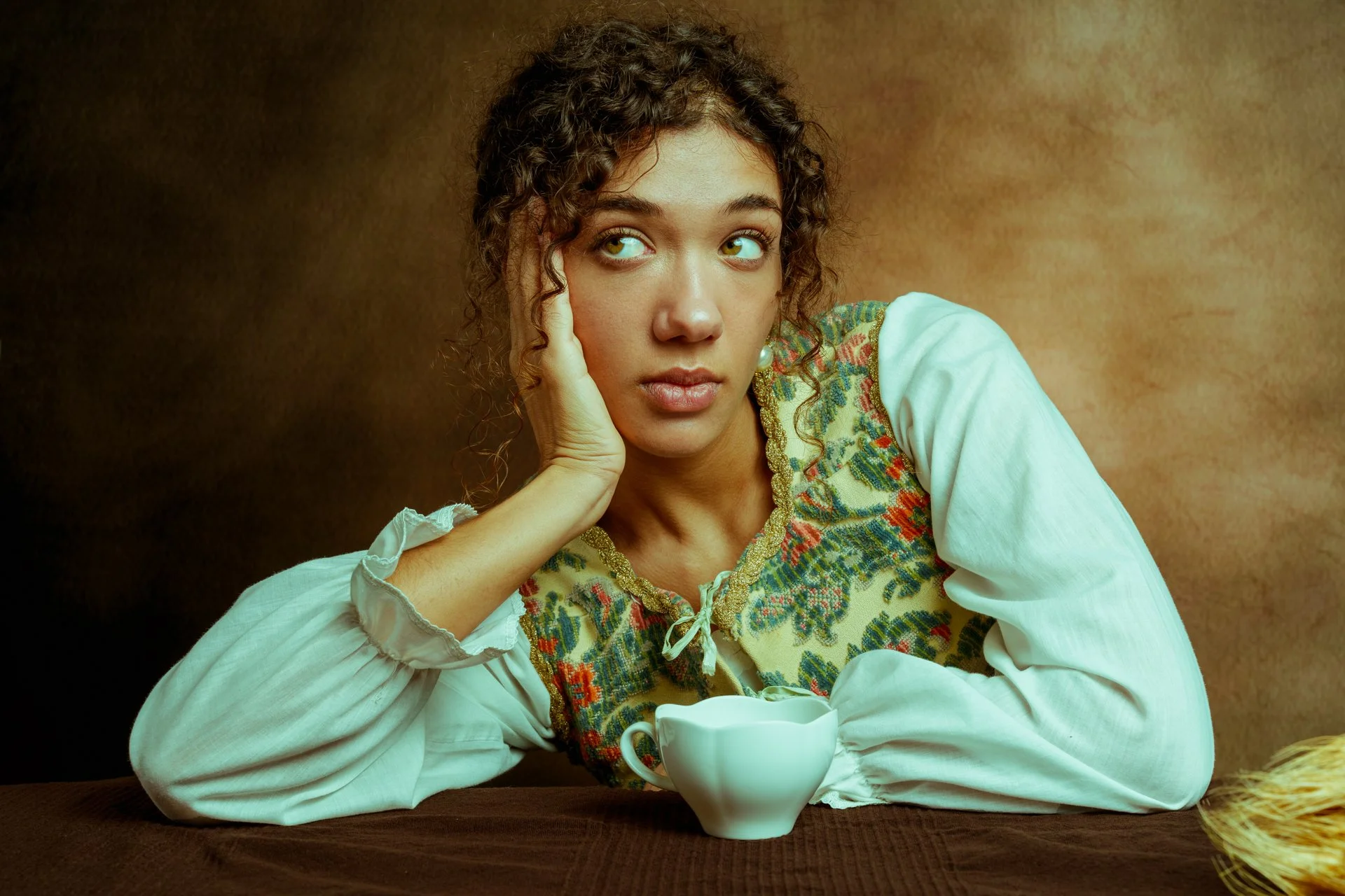 Ragazza seduta con una tazza di ceramica bianca, poggiando il viso sulla mano, sfondo marrone e calmo, abbigliamento con ricami colorati.