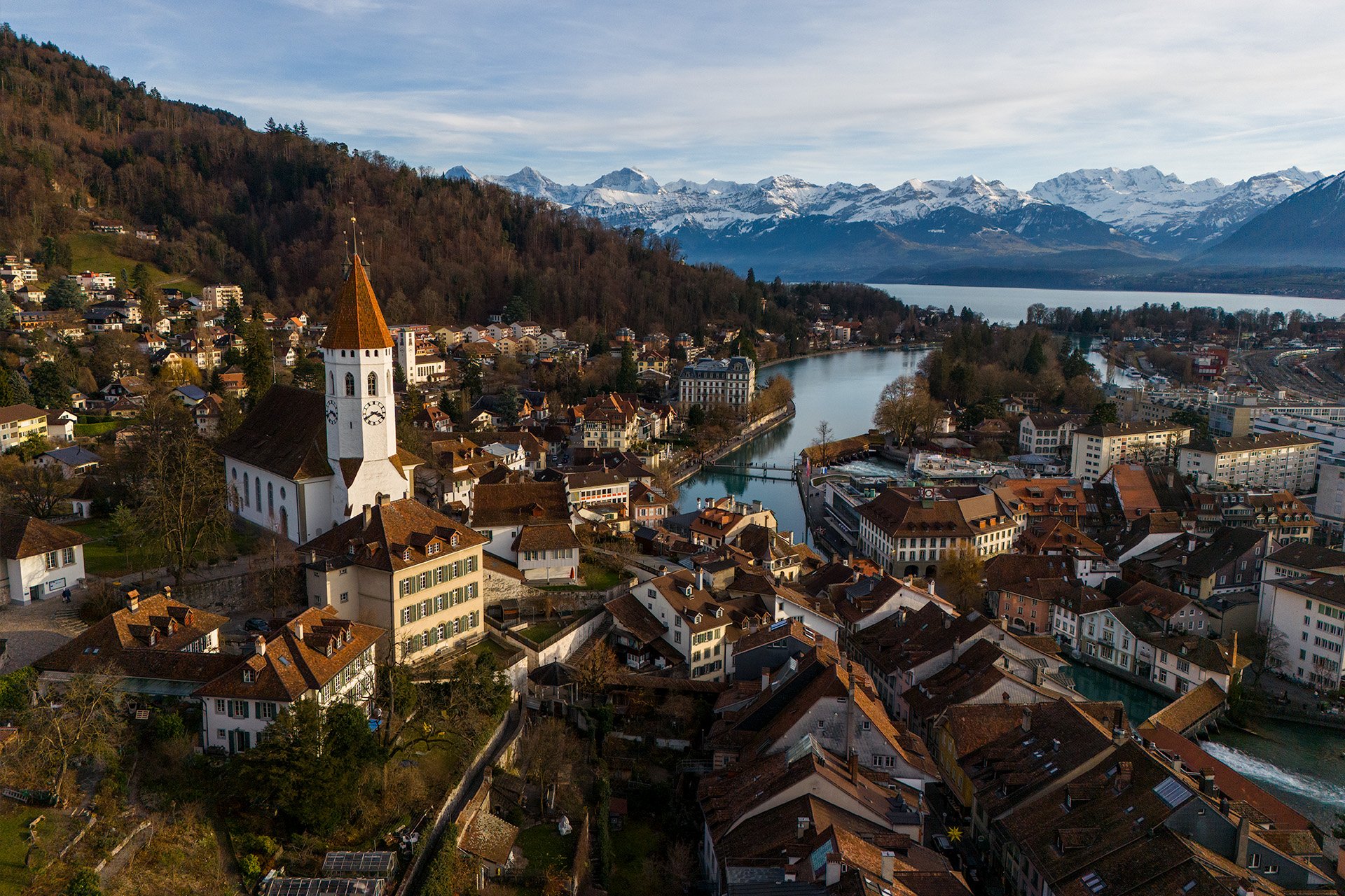 Veduta aerea di una città con una chiesa, case e un lago circondato da montagne innevate.