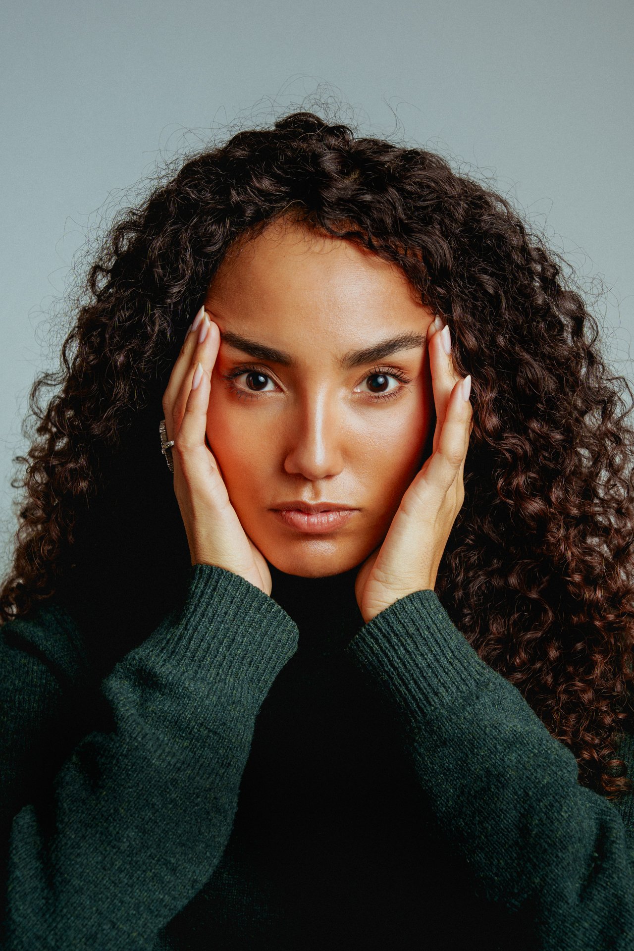 Una donna con capelli ricci, occhi grandi e espressione seria, che si tocca i lati del viso con le mani, indossa un maglione scuro e uno sfondo neutro.