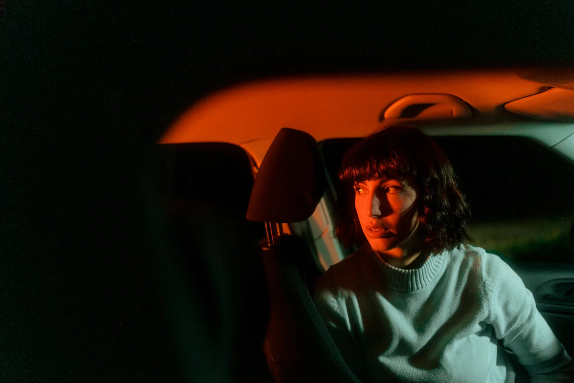Una donna con capelli corti e scuri, vestita con una felpa chiara, seduta nel sedile anteriore di una macchina, illuminata da luci arancioni e verdi, con un'espressione sorpresa o sbalordita.