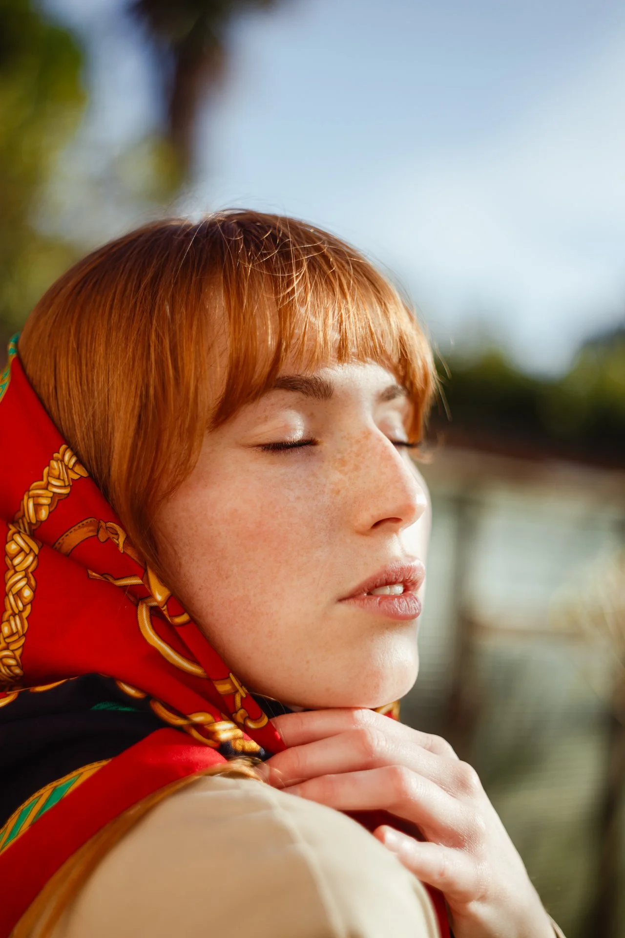 Ragazza con capelli rossi e pelle punteggiata di lentiggini, chiude gli occhi e sorride leggermente, indossa una sciarpa colorata con dettagli dorati.