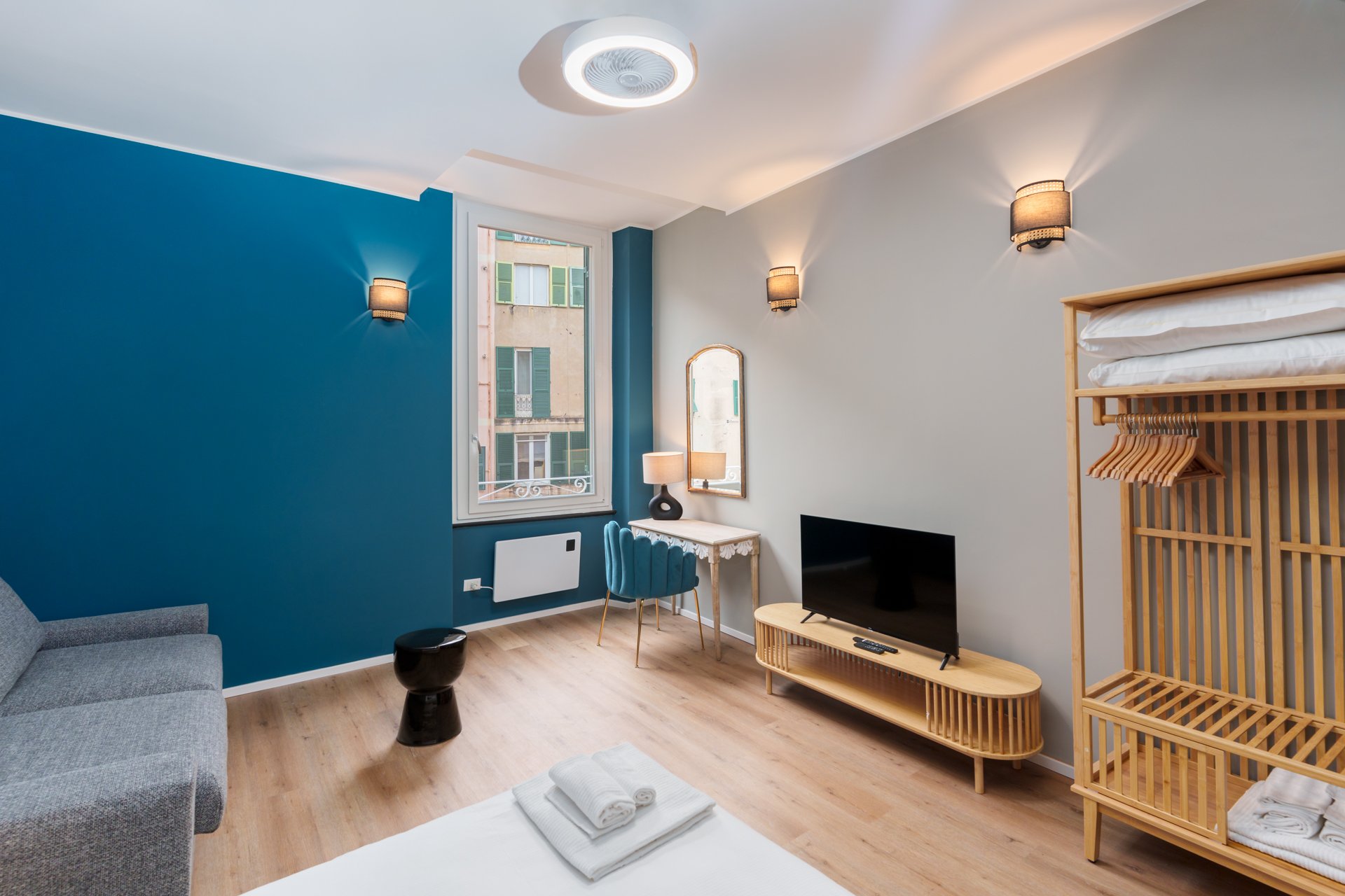 Stanza moderna con pareti bianca e blu, un letto con asciugamani, un divano grigio, un televisore su un mobile in legno, una scrivania con sedia blu, una grande finestra, lampade a parete e un tavolino nero.