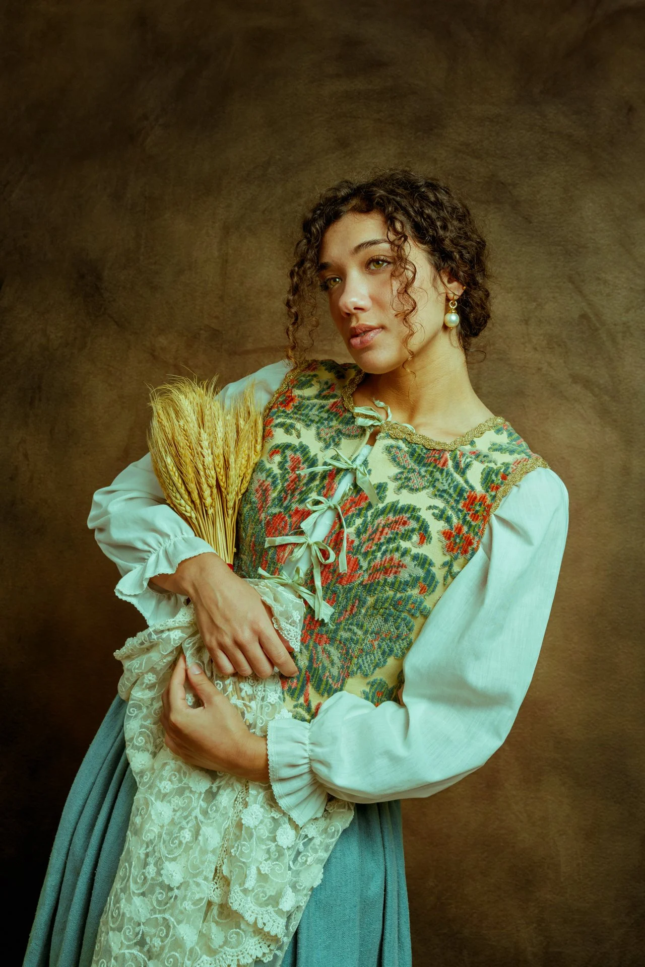 Una donna con capelli riccioli e orecchini di perla, indossa un abito con ricami colorati e una camicia bianca, tiene un mazzo di grano secco e si staglia contro uno sfondo marrone.