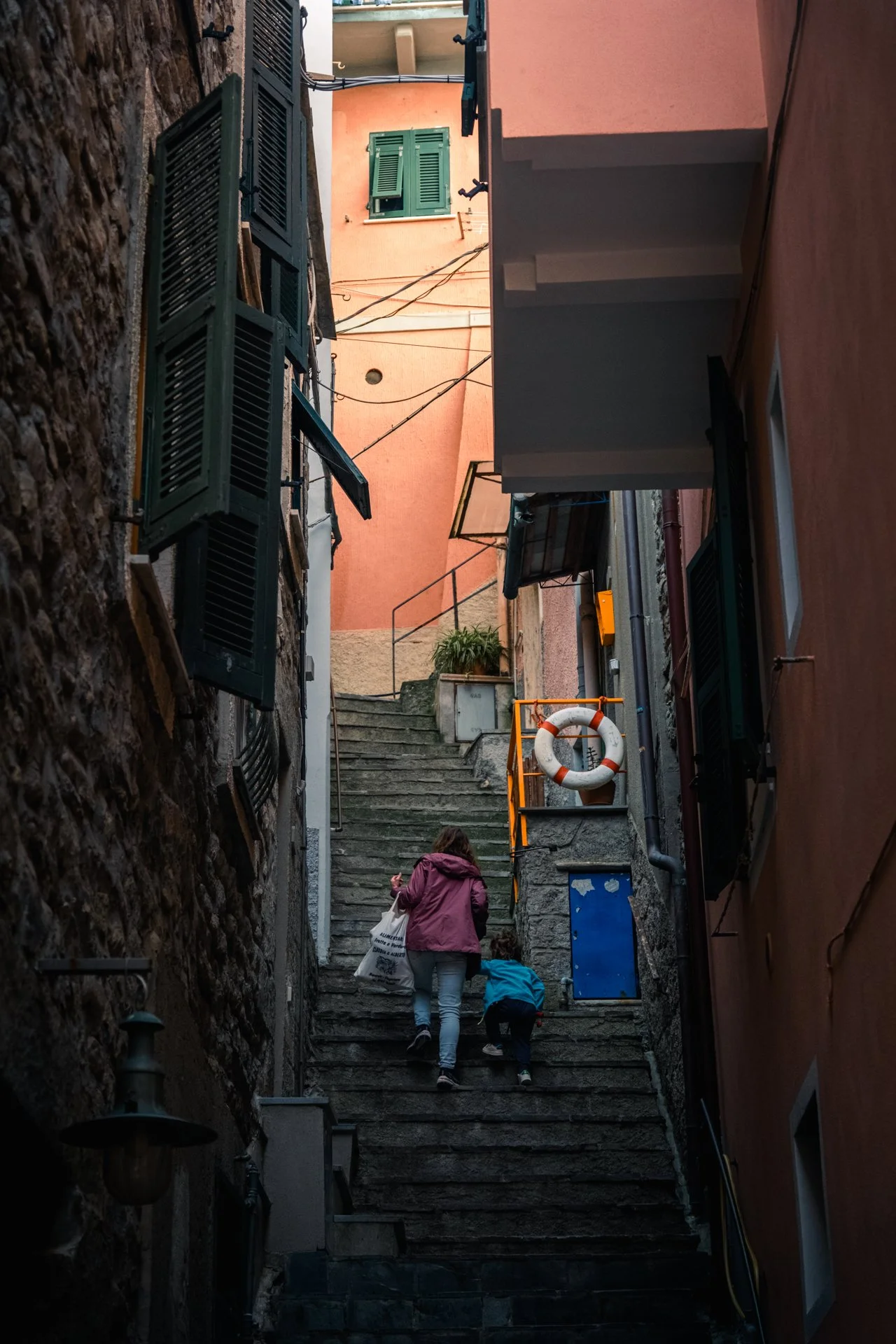 Due persone, una adulta e un bambino, salgono le scale di un vicolo stretto tra edifici con pareti di pietra e colorate, con finestre con persiane e vari oggetti appesi.