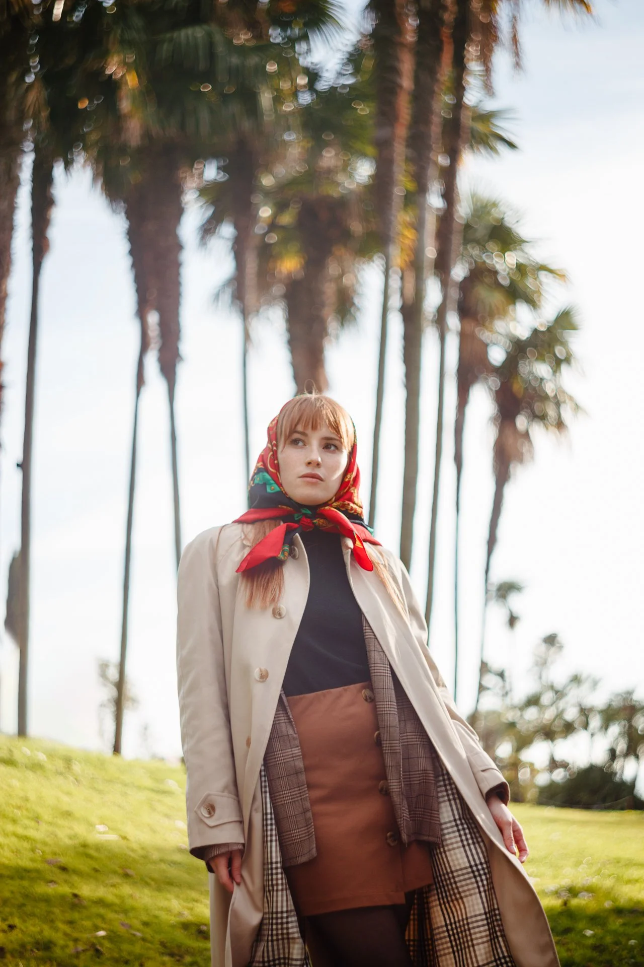 Donna con cappotto beige, gonna marrone, e foulard rossa con motivo floreale, vicino a palme su un tappeto di erba verde.