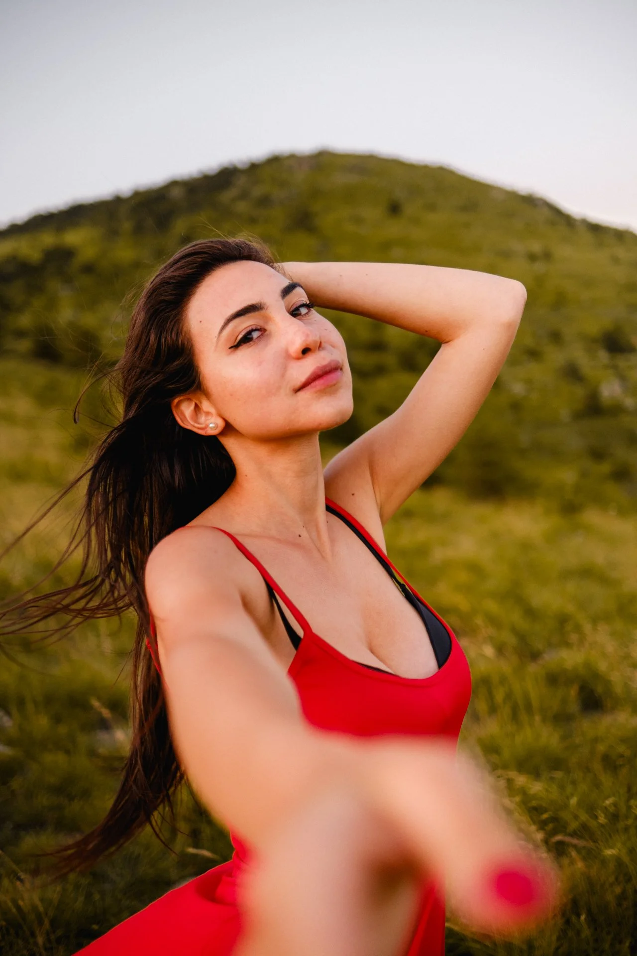 Ragazza con vestito rosso posa in un campo verde con una collina sullo sfondo, scattando una foto in stile selfie.